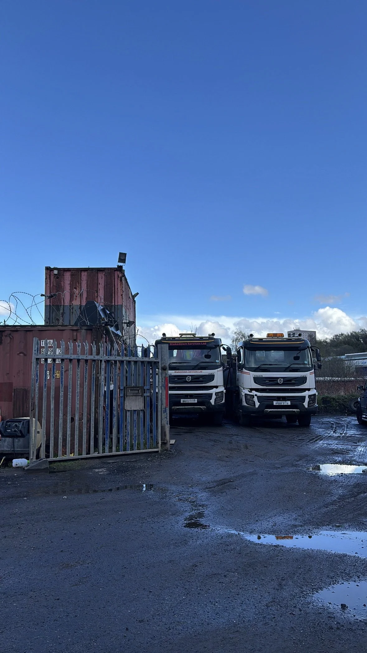 Two white trucks parked behind a metal gate at a facility with a rusted shipping container and barbed wire fence in front. Puddles of water on the ground reflect the sky and trucks. The sky is mostly clear with a few clouds.