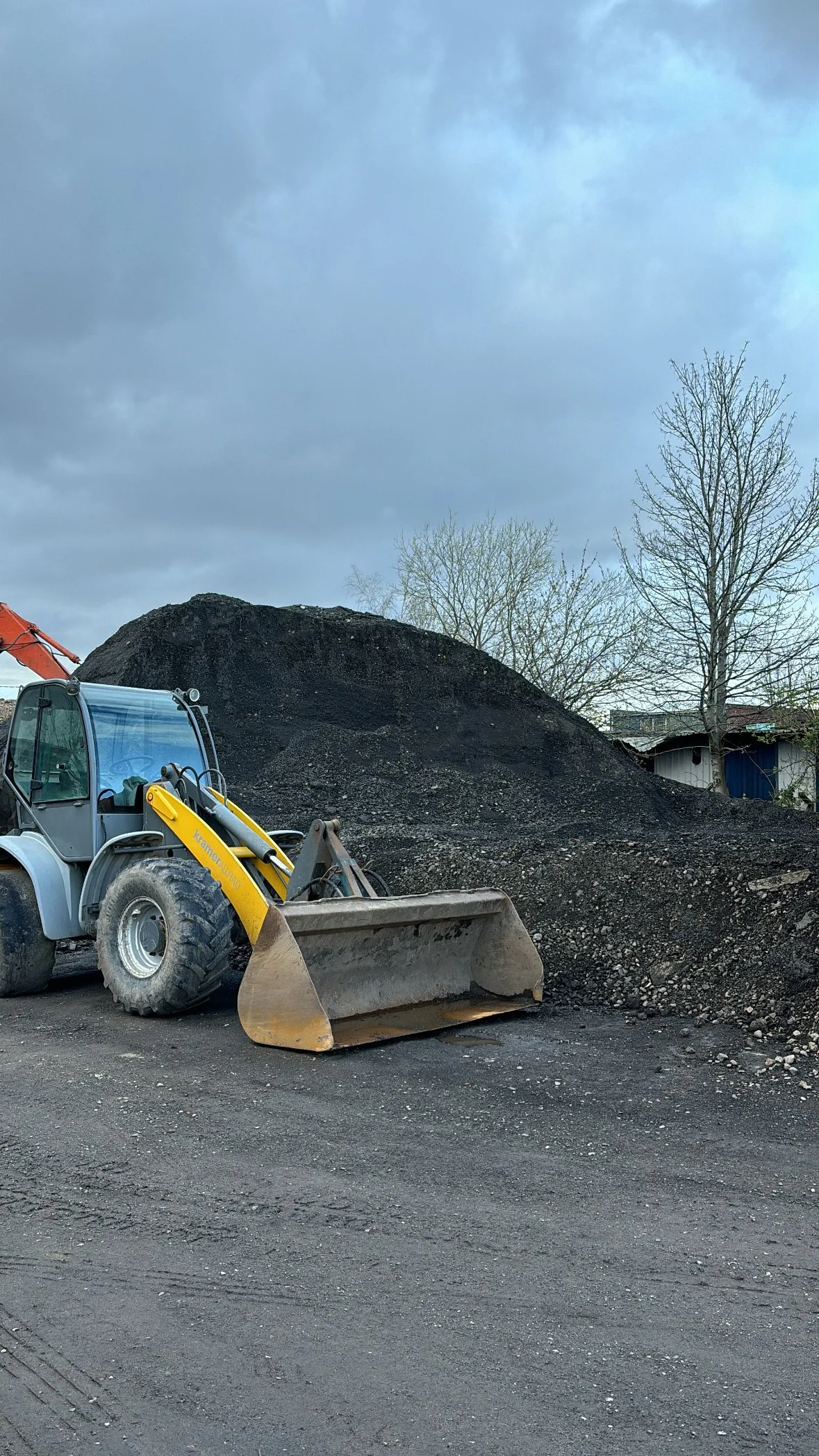 A construction site with a bulldozer in the foreground and a large pile of dirt or gravel in the background, with trees and a partly cloudy sky.