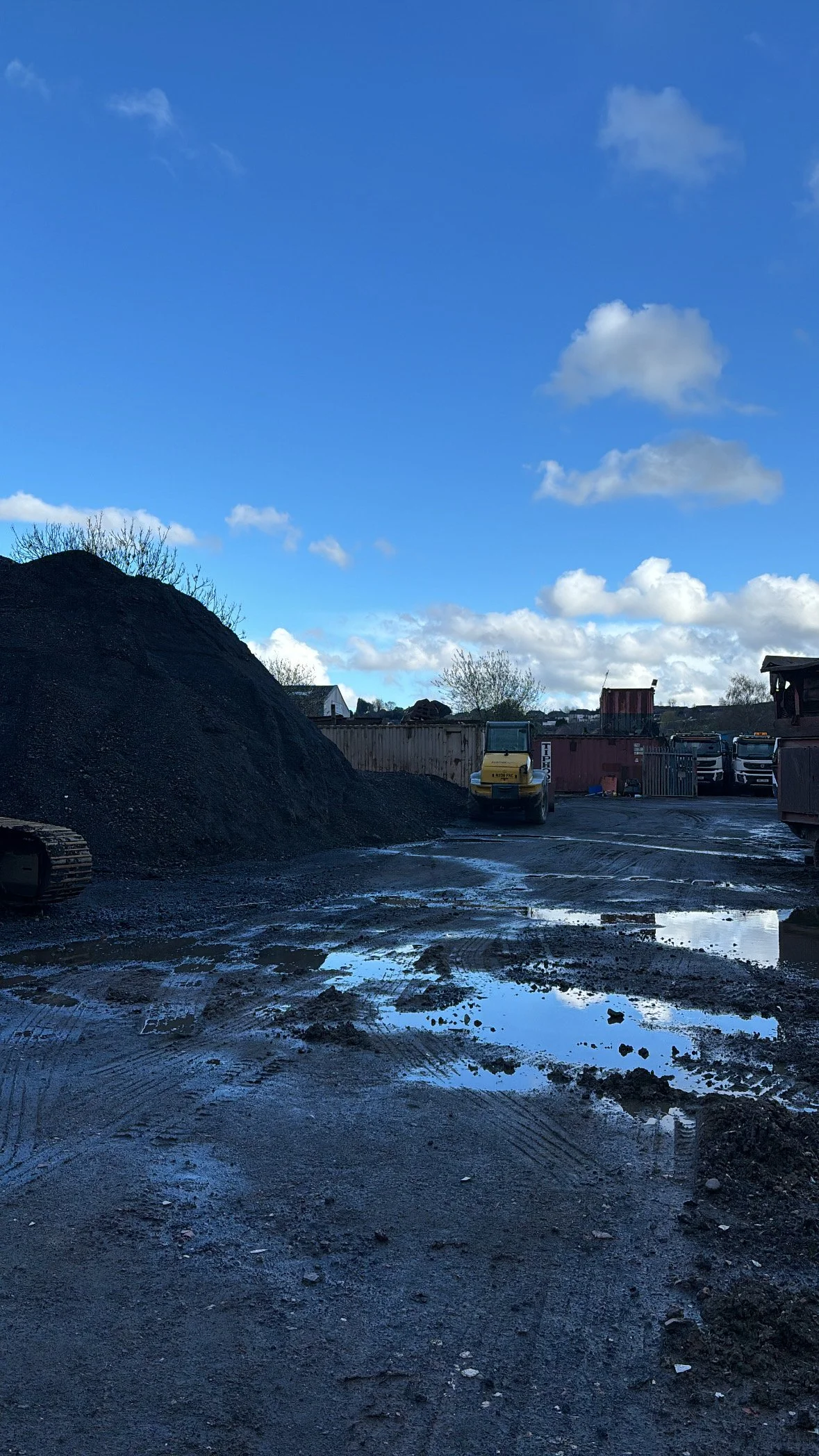 View of a construction site with a pile of gravel on the left, muddy ground with puddles, and construction vehicles including a yellow trailer and trucks in the background under a blue sky with scattered clouds.