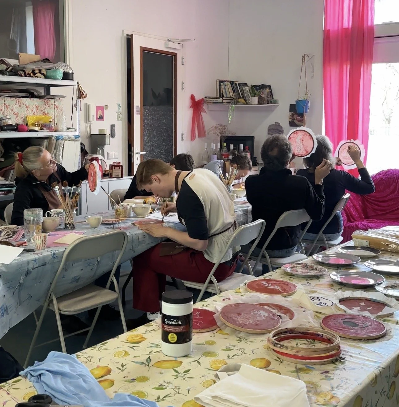 People sitting around a table painting ceramic plates with pink designs, in a well-lit room with pink curtains and decorated walls.