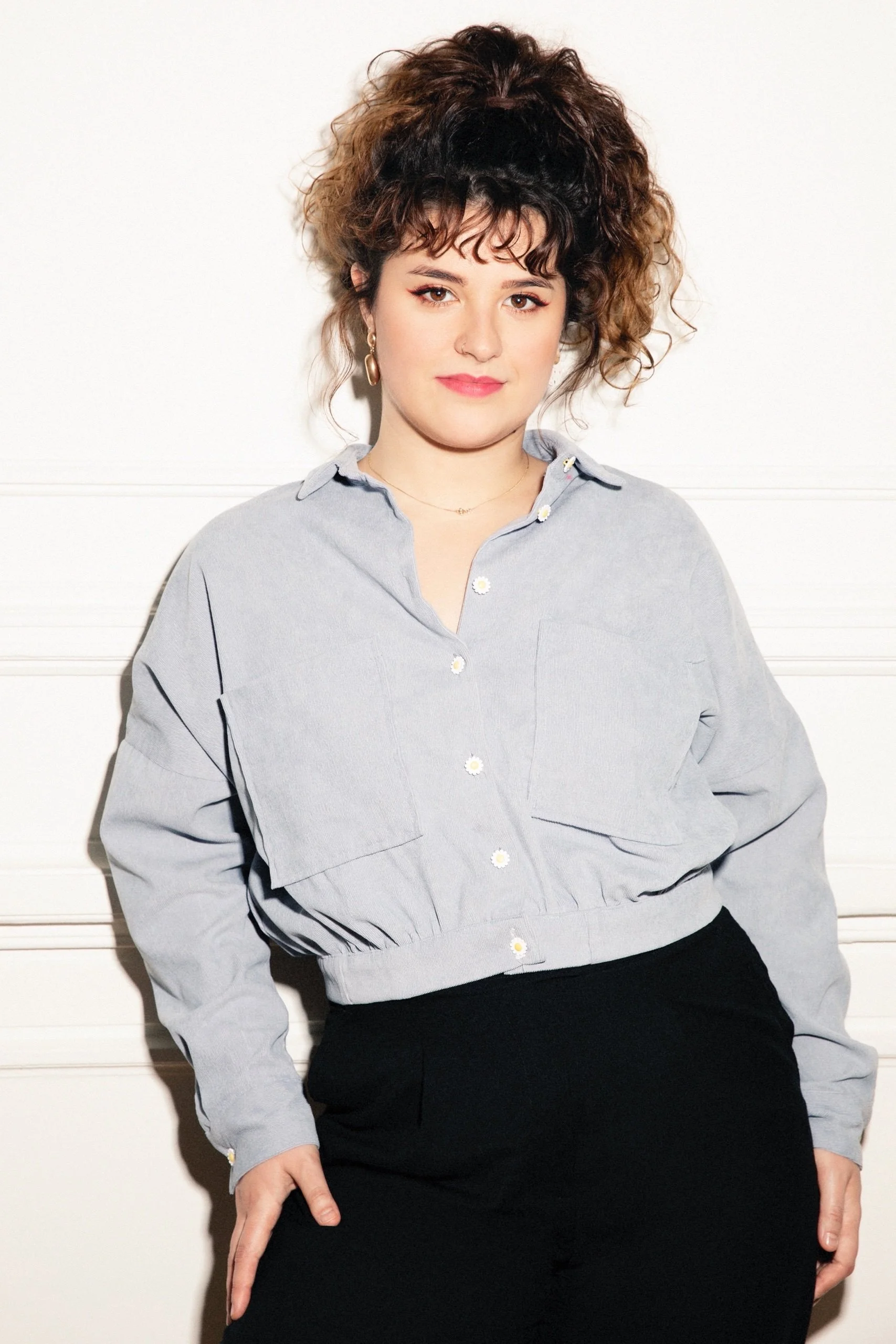 A woman with curly brown hair, wearing a light gray button-up shirt with daisy details, earrings, and a necklace, stands against a white background.