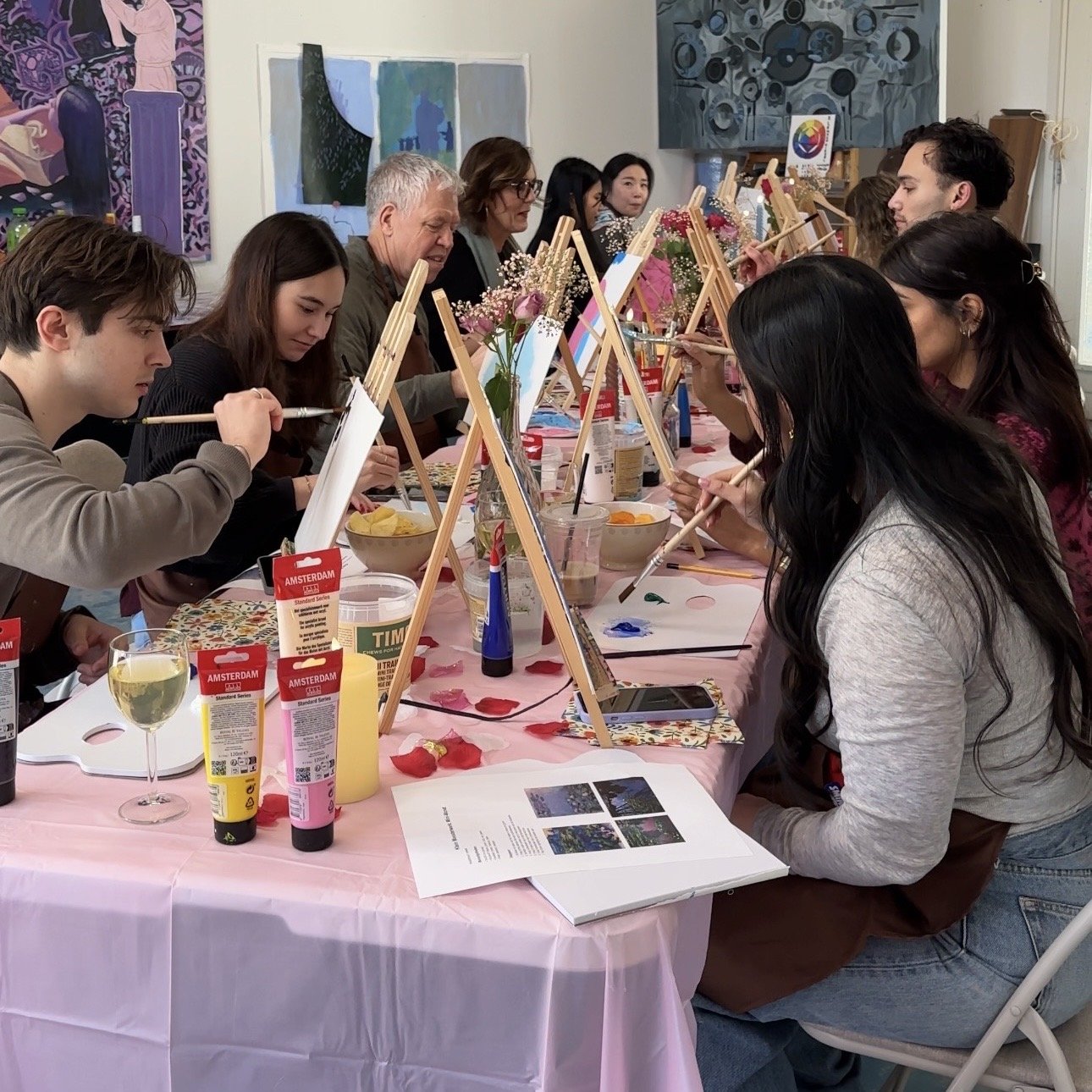 A group of people participating in a group painting class, sitting at a pink-covered table with easels, brushes, and paint supplies, with artwork displayed on the walls behind them.