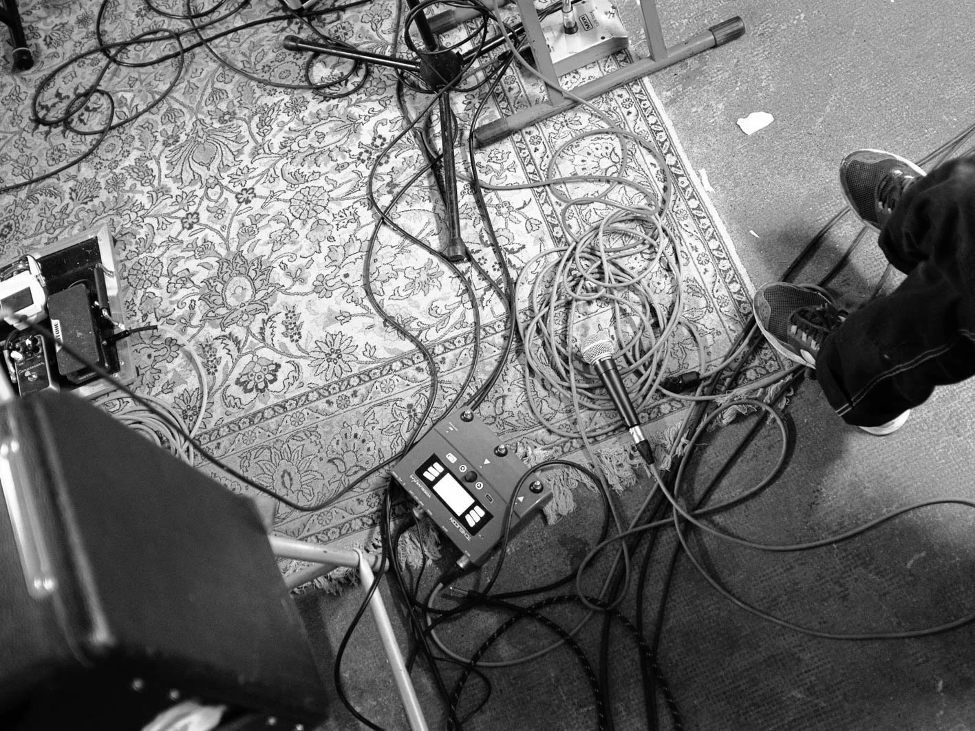Câble audio, microphone et équipements de sonorisation disposés sur un tapis et un sol en dur, avec deux personnes debout à proximité, probablement une scène ou un studio d'enregistrement.