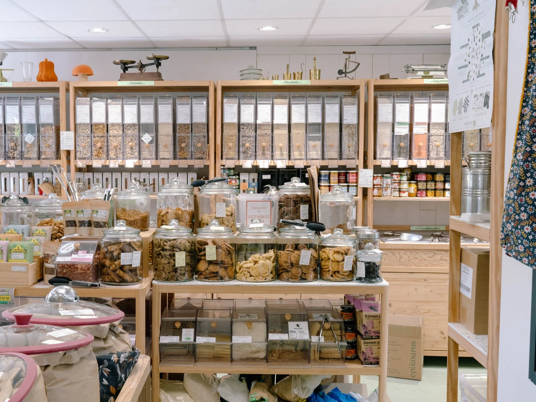 Rayons de produits en vrac dans une épicerie proposant des céréales, biscottes et biscuits, avec des bocaux en verre contenant des produits secs. Structure en bois avec étiquettes indiquant les catégories.