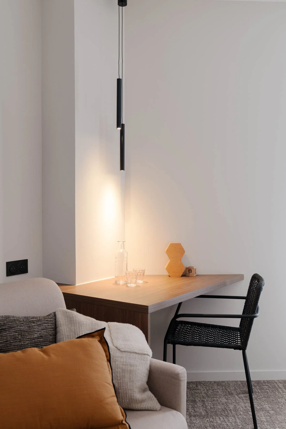 Un coin salon avec un canapé beige, coussins gris et marron, un bureau en bois avec une bouteille en verre, deux verres, un objet en forme d'hexagone en bois, une petite boîte, et une chaise noir.