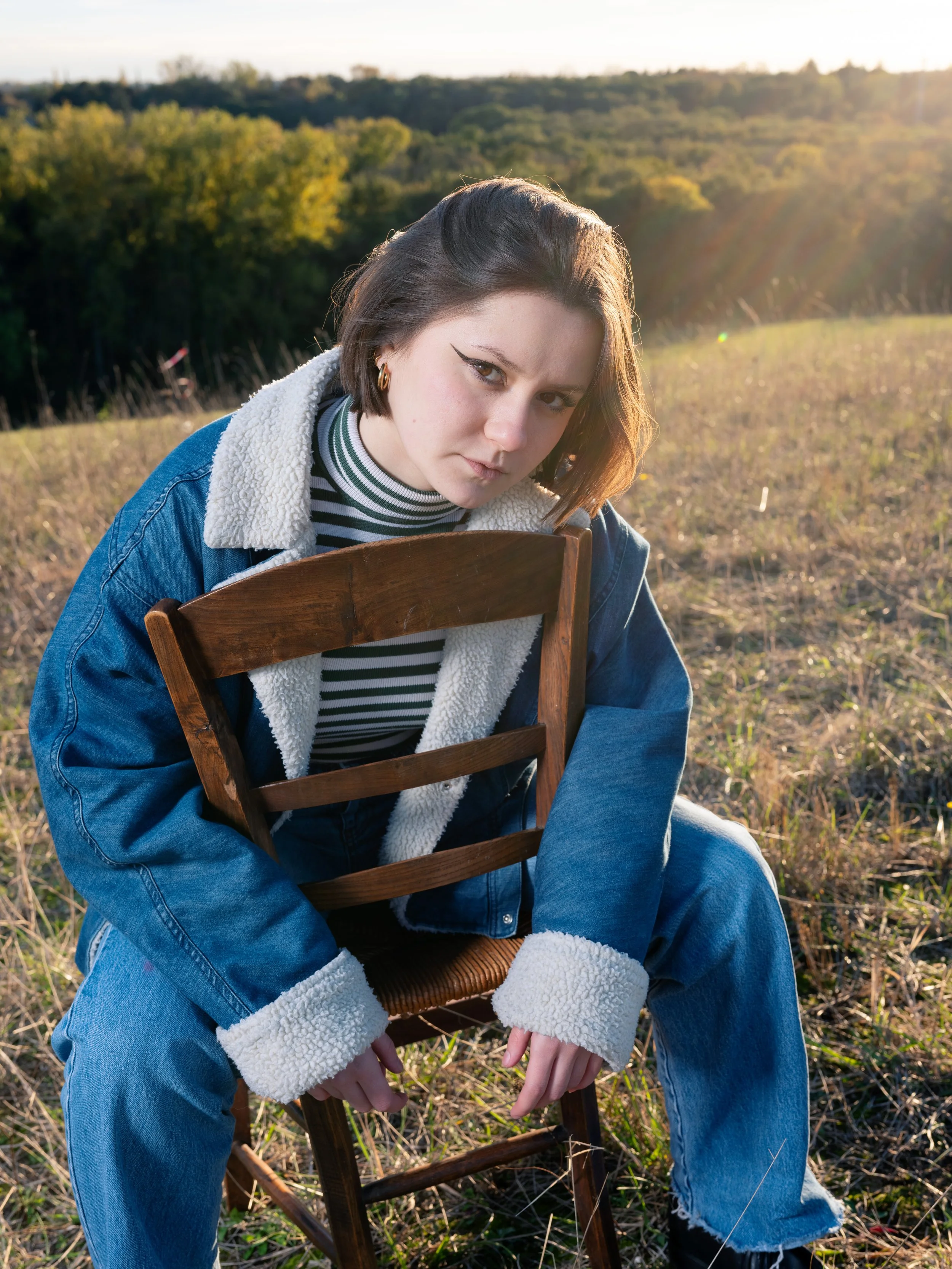 Une jeune femme avec des cheveux bruns courts, portant un pull rayé noir et blanc, une veste en jean avec une doublure en peluche, et des boucles d'oreilles, assise en extérieur dans un champ avec des arbres en arrière-plan, au coucher du soleil.