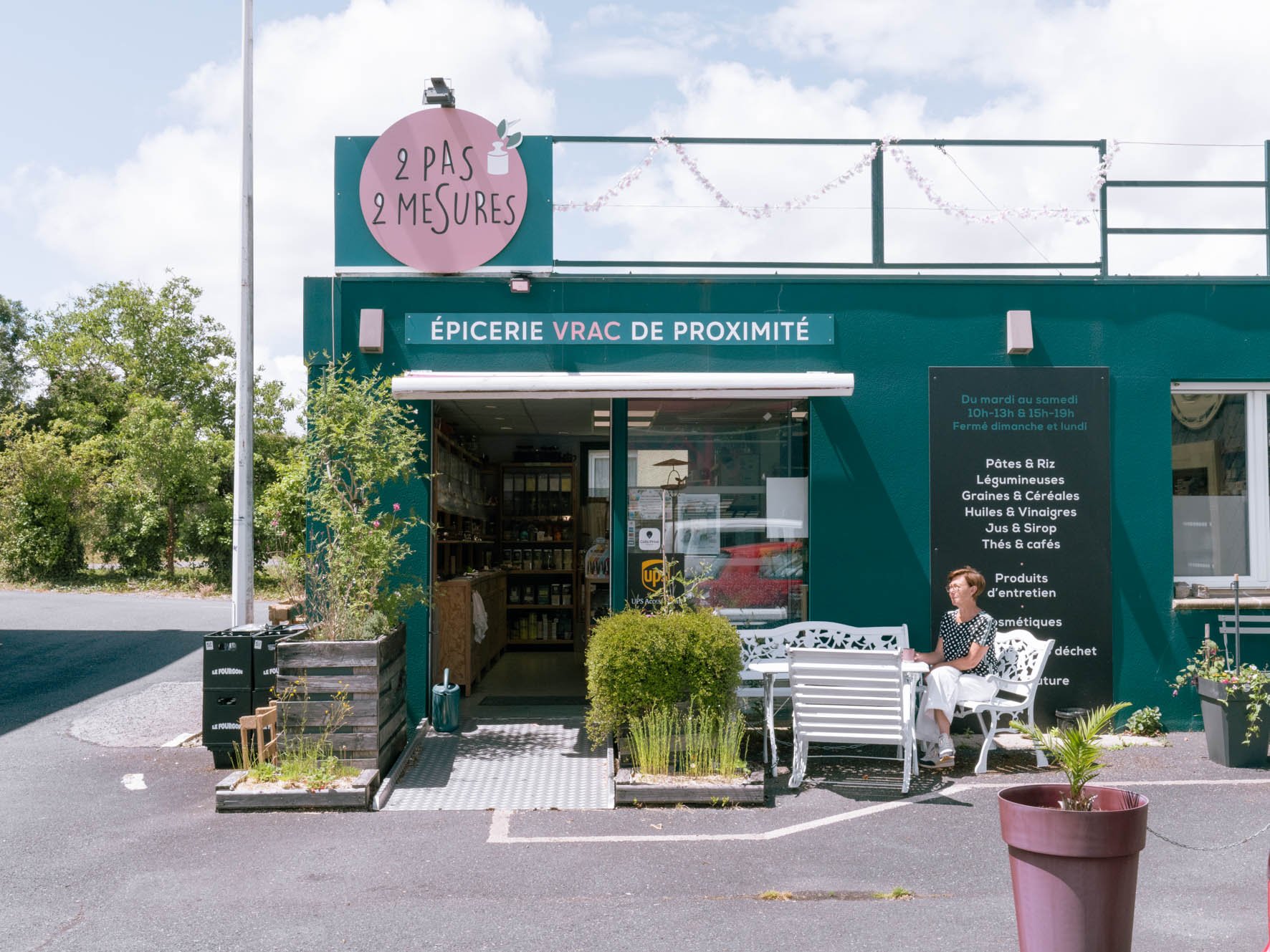 Façade d'une épicerie en vrac de proximité avec une terrasse où une femme est assise sur un banc blanc. Le magasin affiche un panneau indiquant ses horaires et une liste de produits proposés tels que pâtes, riz, légumes, graines, huiles, vinaigres, jus, sirops, thés, cafés, produits d'entretien, cosmétiques, et déchets.