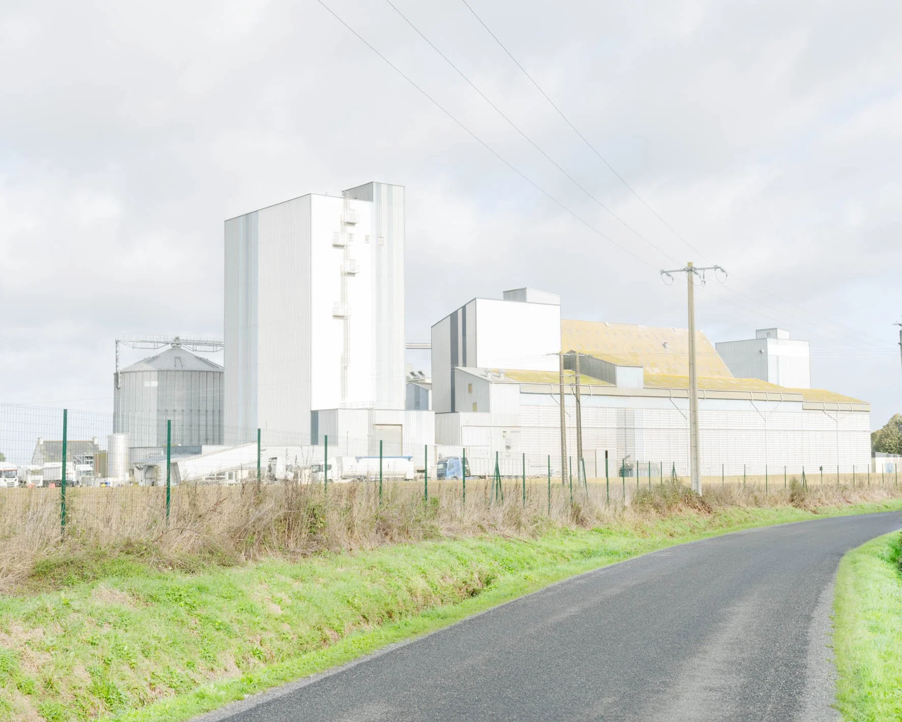 Bâtiments industriels blancs avec toits en tôle, terrain entouré d'une clôture en fil de fer vert, chemin asphalté courbe, poteaux électriques, ciel nuageux.
