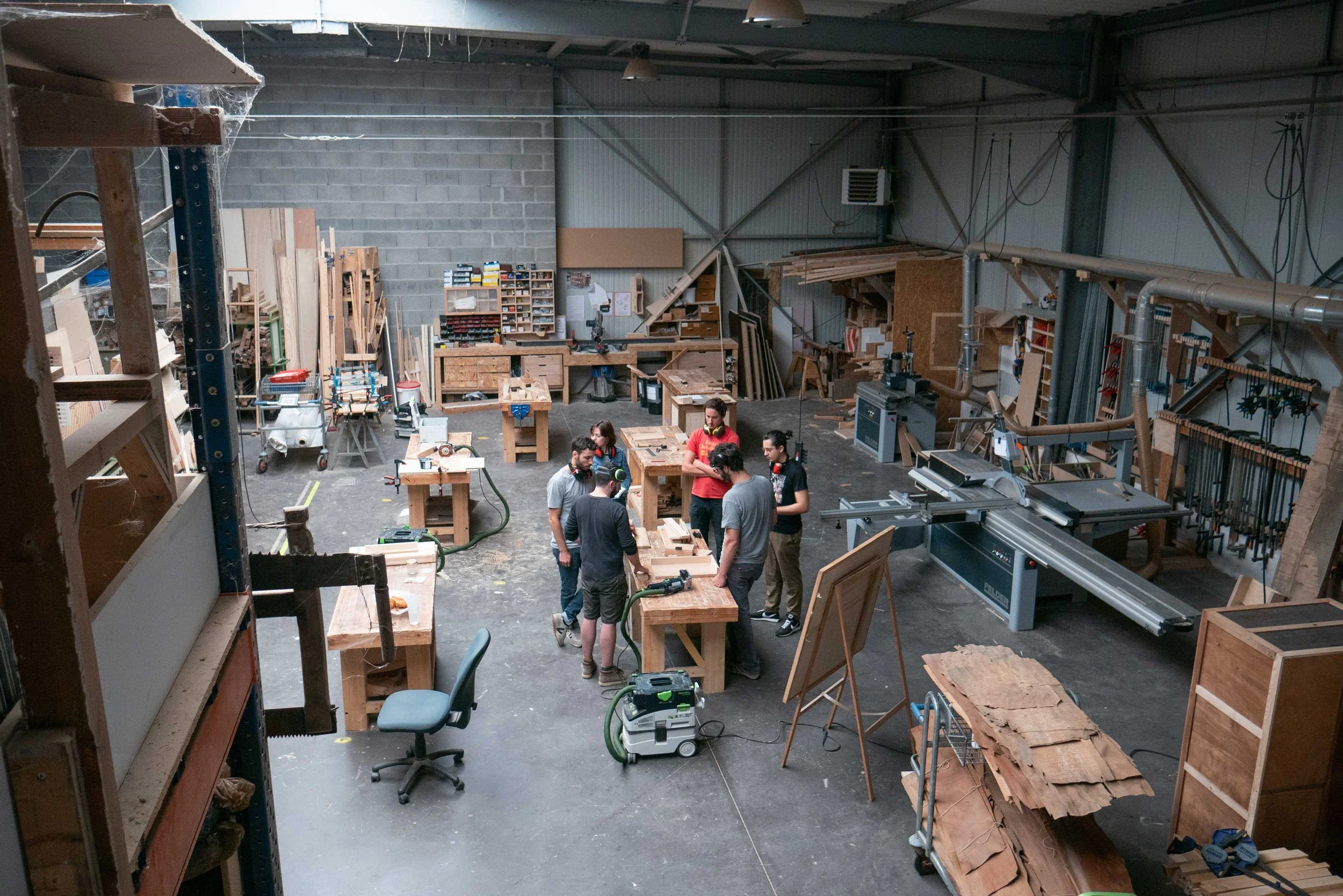 Atelier de menuiserie avec plusieurs personnes travaillant sur des plans en bois et des machines dans un environnement de bricolage. Des étagères en bois, des outils, des chaises et du matériel divers.
