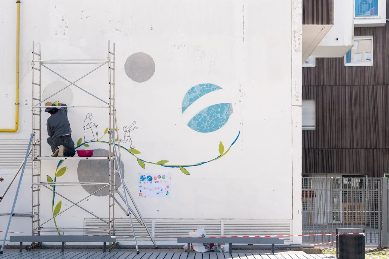 Un artiste peint un mural sur un mur blanc, utilisant une échelle en métal. Le mural comporte des éléments abstraits et des dessins de deux enfants. Le décor environnant est urbain avec un bâtiment en bois et en béton, ainsi que des zones clôturées.