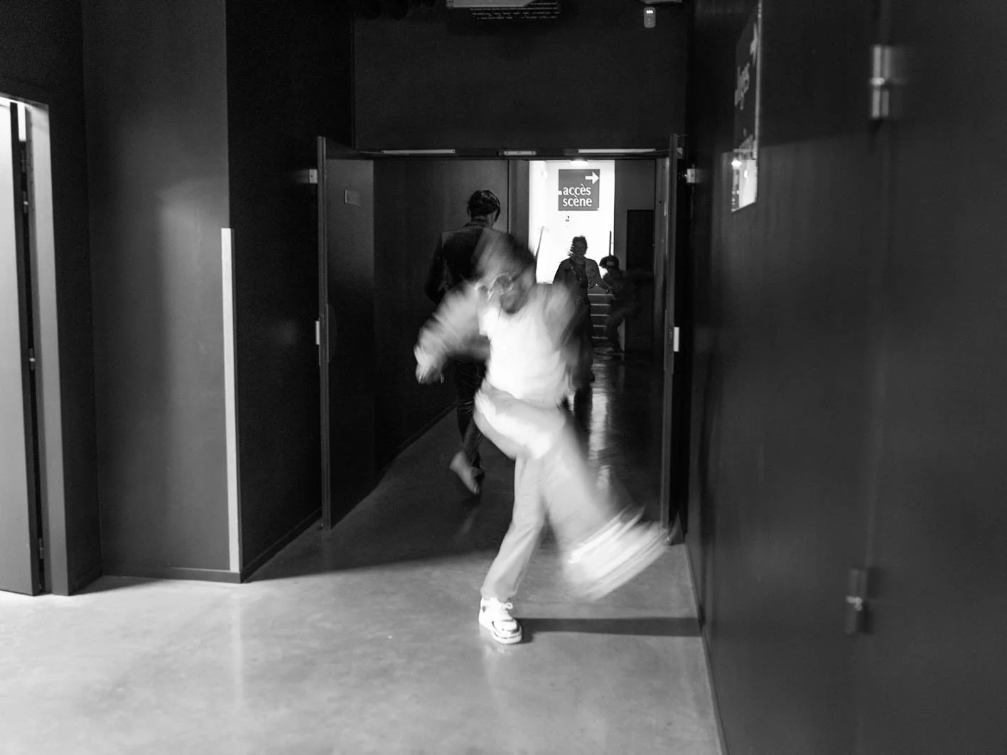 Des enfants courent dans un couloir sombre, une porte entrouverte au fond avec un panneau 'accès scène', l'image est en noir et blanc.