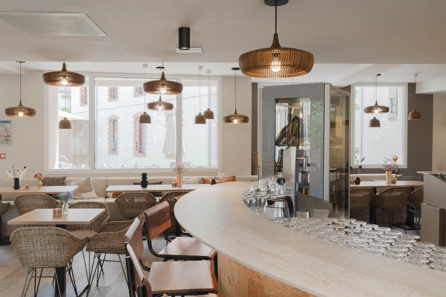 Intérieur d'un café ou restaurant avec des tables, chaises en rotin, décorations florales, un bar avec verres alignés, et des lampes suspendues en bois.