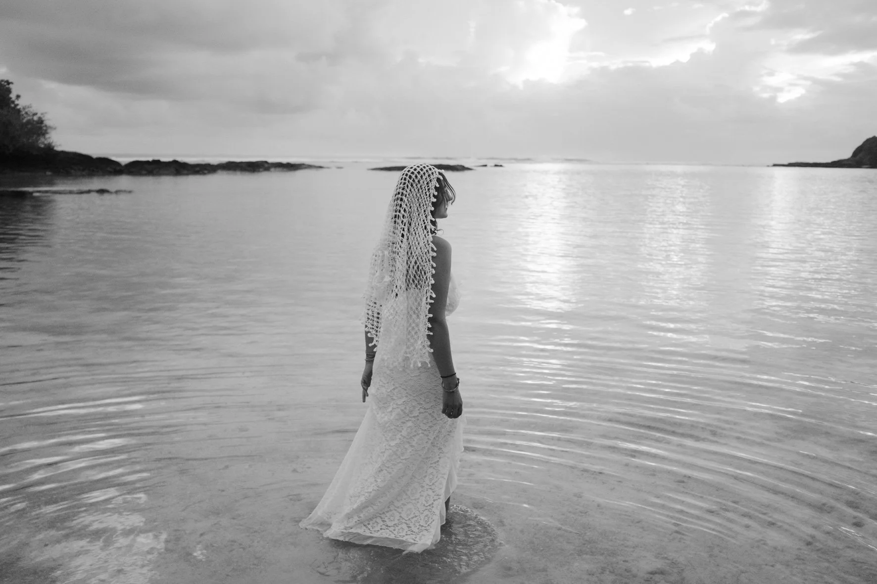 Une femme en robe de mariée tient la main dans l'eau à la plage, portant un voile crocheté, avec un ciel nuageux et une mer calme en arrière-plan.