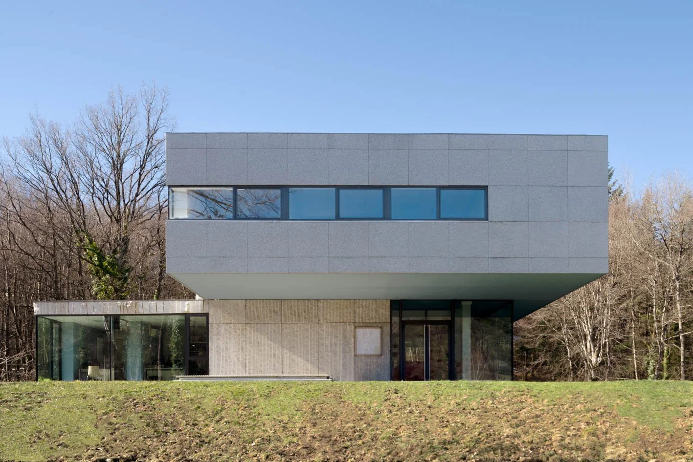 Une maison moderne en béton avec deux niveaux, grandes fenêtres, en milieu naturel avec des arbres en arrière-plan et une pelouse devant.