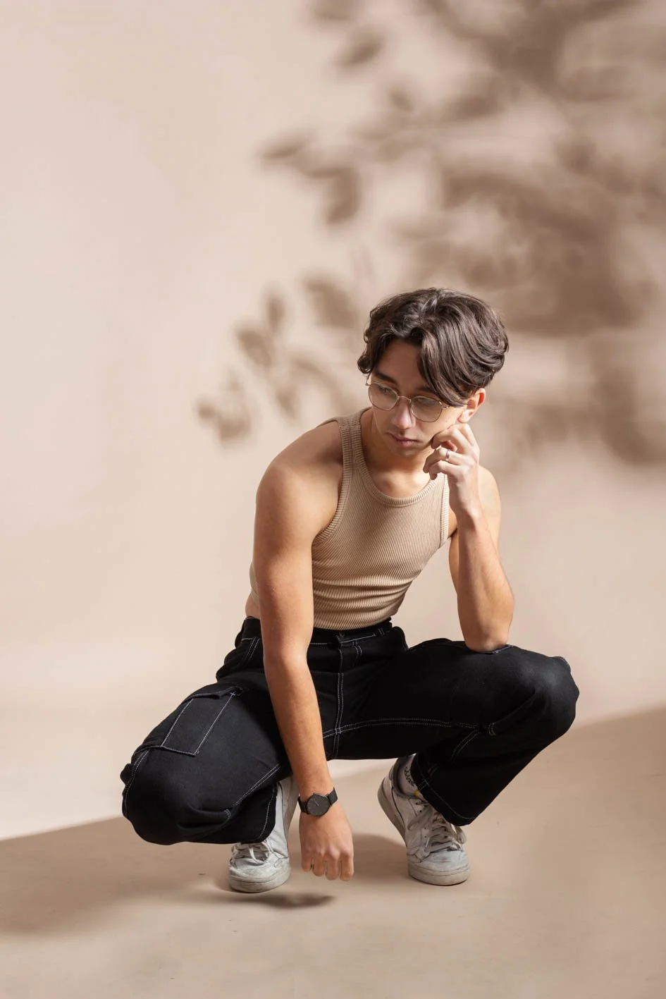 Un jeune homme aux cheveux bruns, portant lunettes, une rasgée beige, un pantalon noir et des baskets blanches, est en position accroupie sur un sol beige devant un fond uni avec un motif flou d'ombre.
