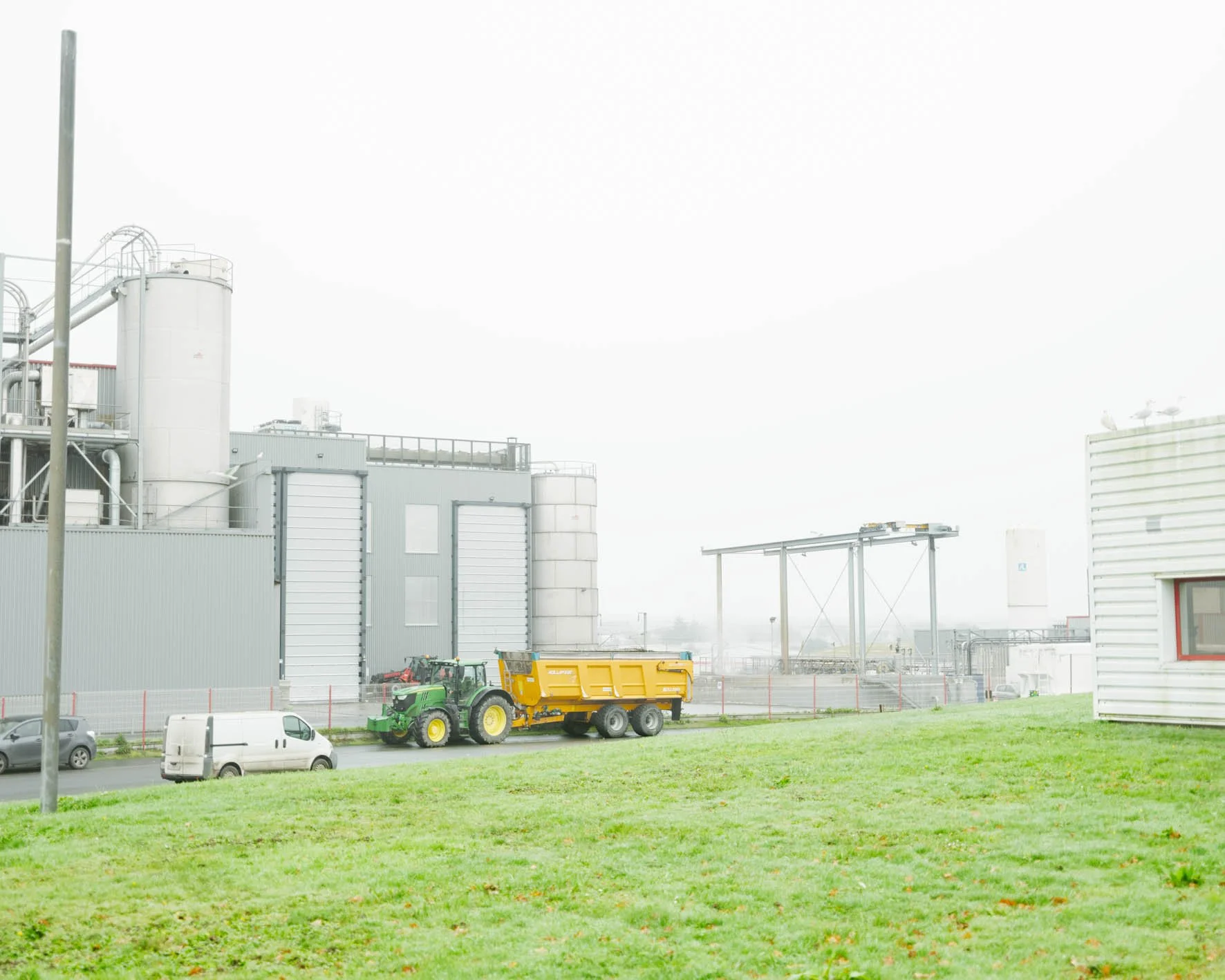 Immeubles industriels avec silos, une pelouse verte au premier plan, et un tracteur jaune de couleur vert, ainsi que des voitures en arrière-plan.