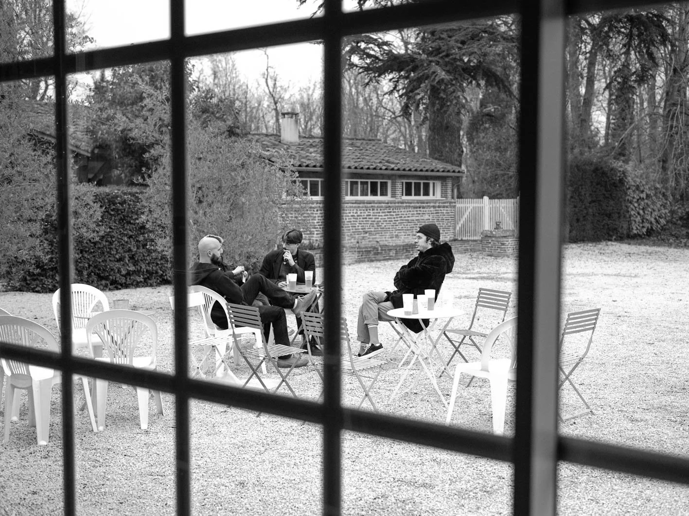 Trois personnes assises dehors, en train de discuter, vues à travers une grille. Il y a des tables avec des boissons sur une terrasse en gravier, avec un bâtiment et des arbres en arrière-plan.