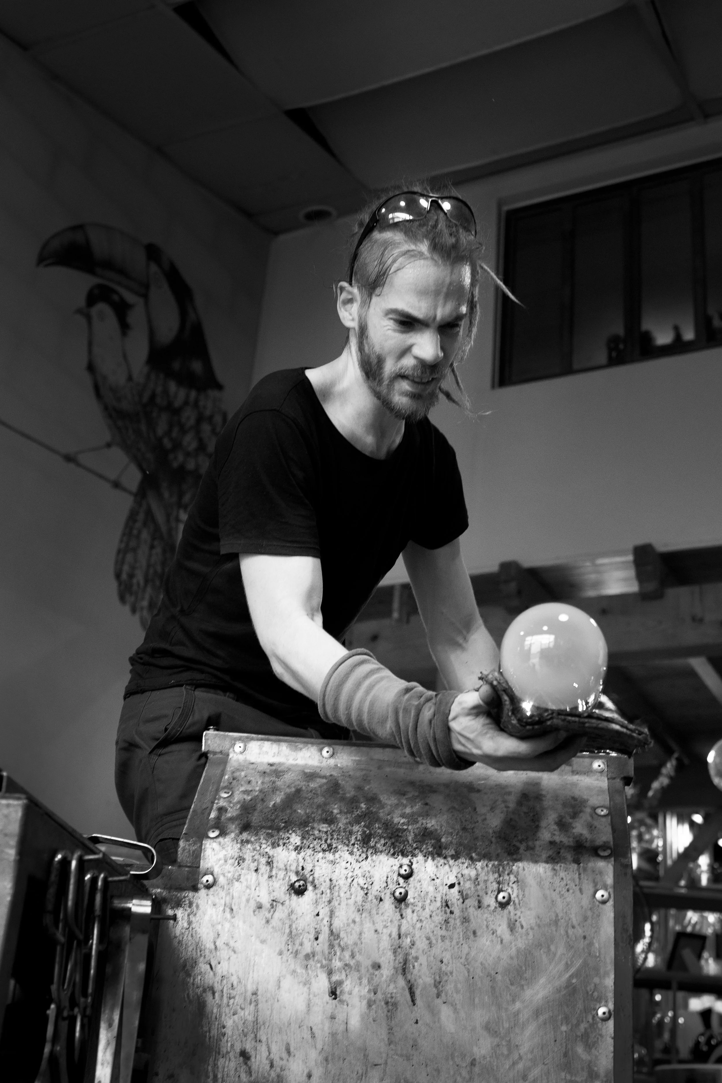 Un homme black et blanc avec des dreadlocks, portant des lunettes sur la tête, travaille avec un objet en verre lumineux dans une galerie ou un studio, derrière un comptoir de travail.