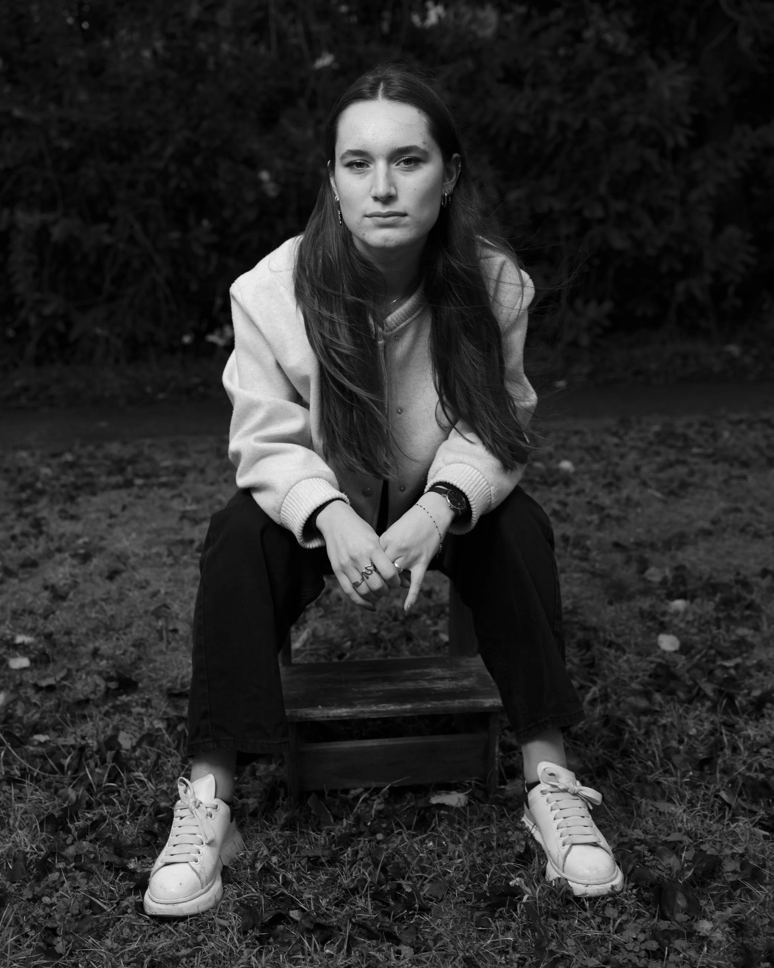Une jeune femme assise sur une chaise en plein air, portant un pull clair, des bijoux, et des baskets blanches, avec un fond d'arbustes et de feuilles mortes au sol. L'image est en noir et blanc.