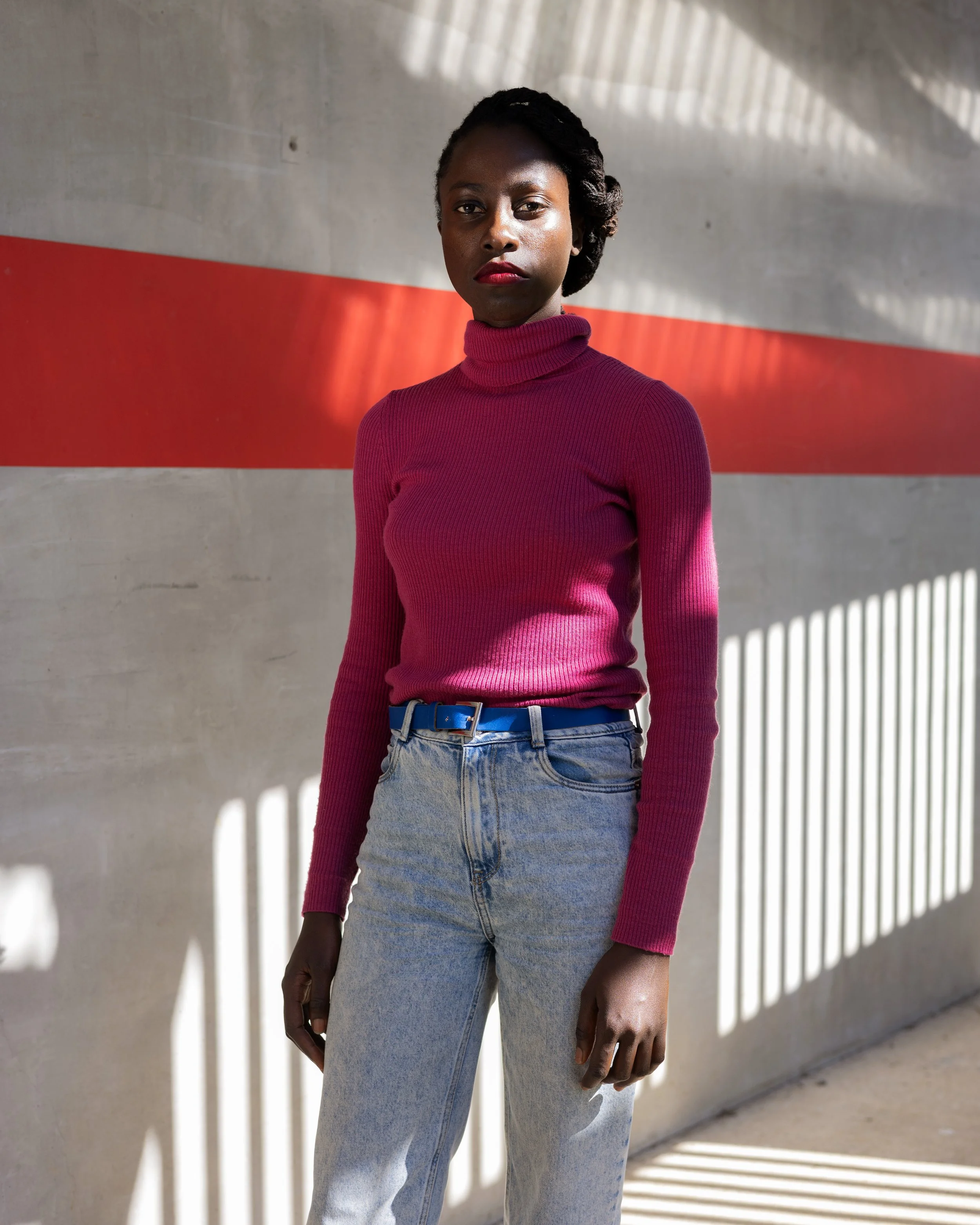 Une femme debout contre un mur en béton, portant un pull ras du cou rose foncé et un jean clair, avec des rayons de lumière créant des ombres sur le mur.
