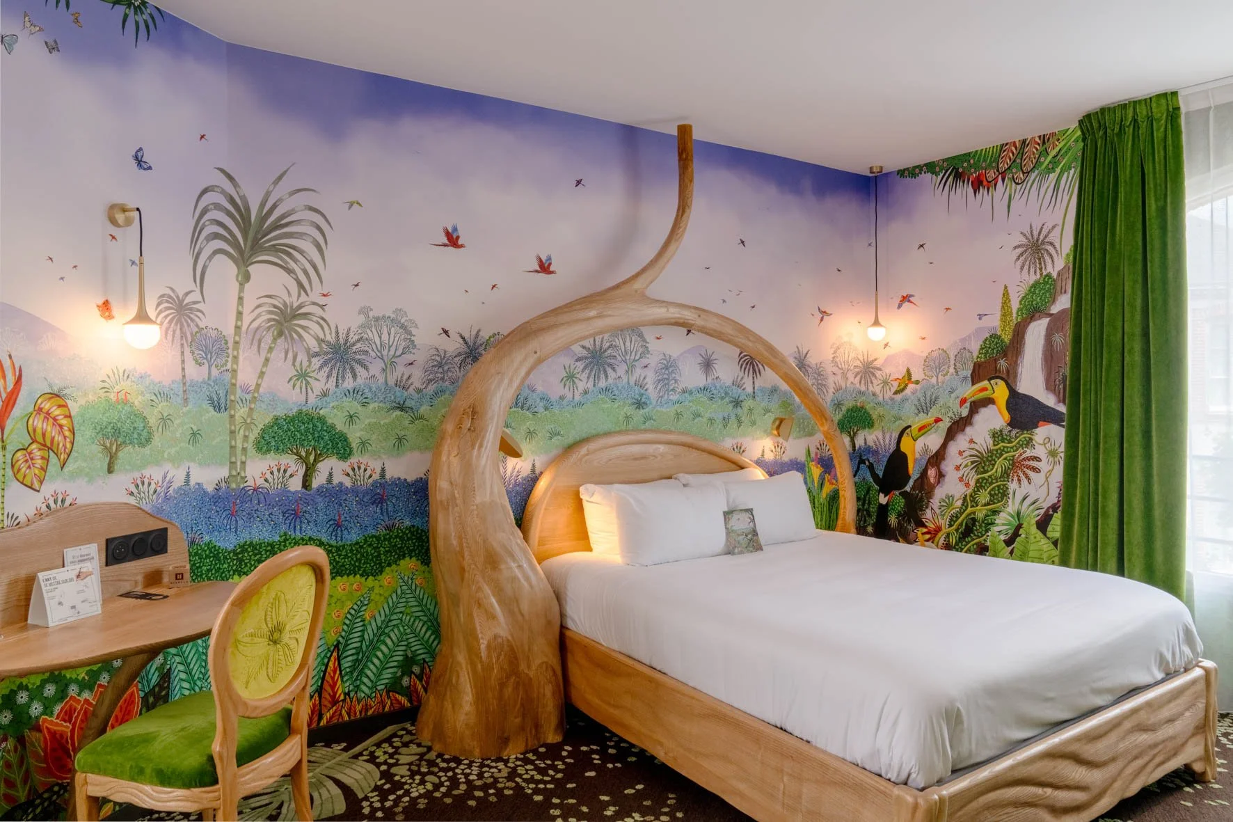 Chambre d'hôtel avec mur peint représentant une forêt tropicale, des oiseaux, des singes et une cascade, avec un lit en bois sculpté et des rideaux verts.