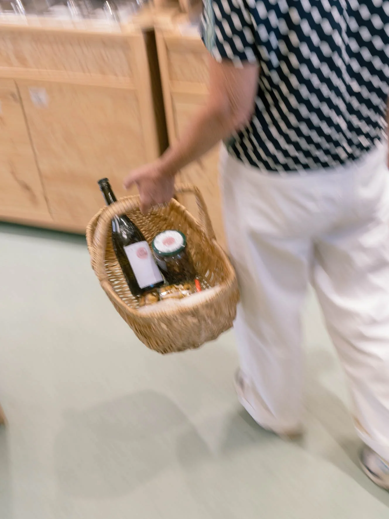 Une personne portant un pantalon blanc et une chemise à motifs noirs et blancs, tenant un panier en osier contenant une bouteille et un pot.