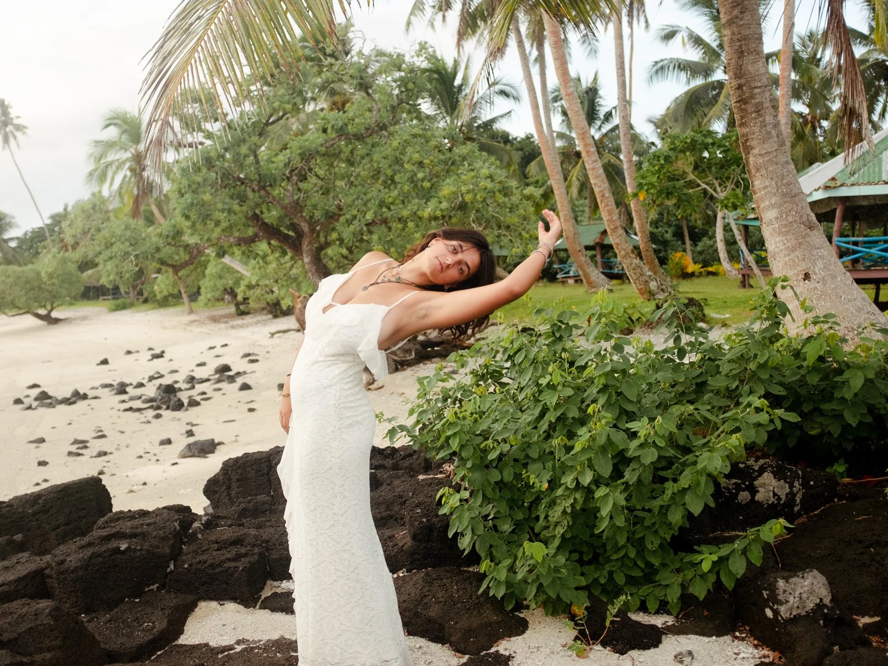 Femme en robe blanche debout sur une plage avec des arbres et des palmiers en arrière-plan, devant des rochers volcaniques et des végétation luxuriante.
