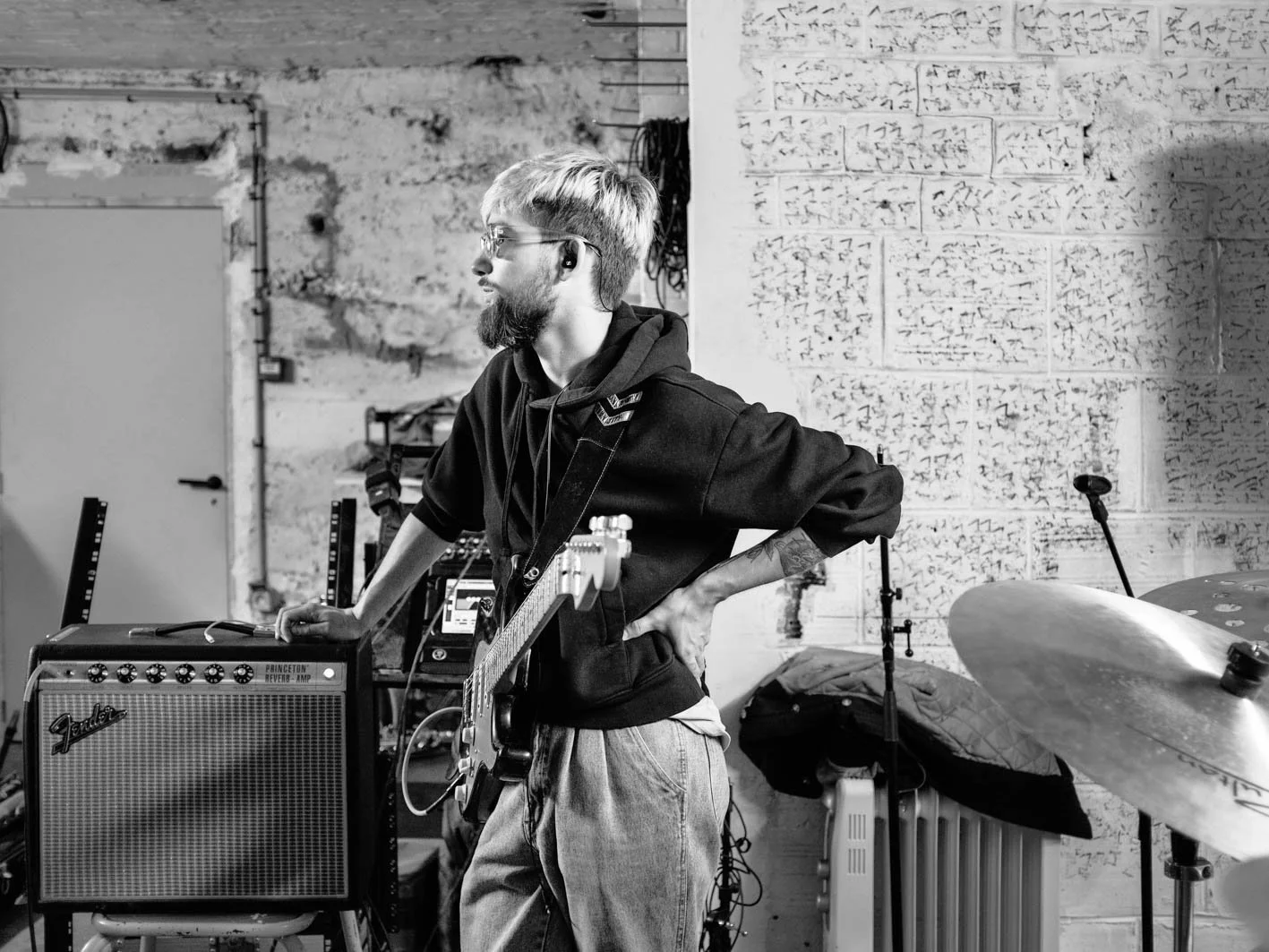 Musicien de profil dans un studio, avec un ampli guitare Fender et un clavier derrière lui, garage ou atelier avec murs en béton.