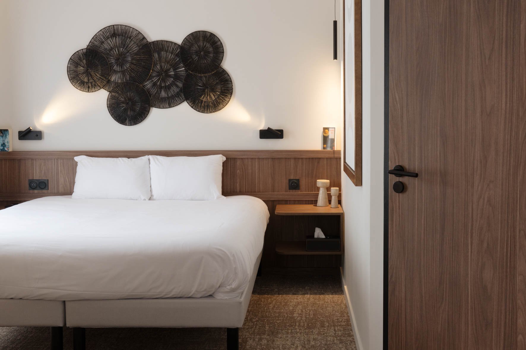 Chambre d'hôtel avec lit blanc, décoration murale en rotin noir, table de nuit en bois, porte en bois derrière.