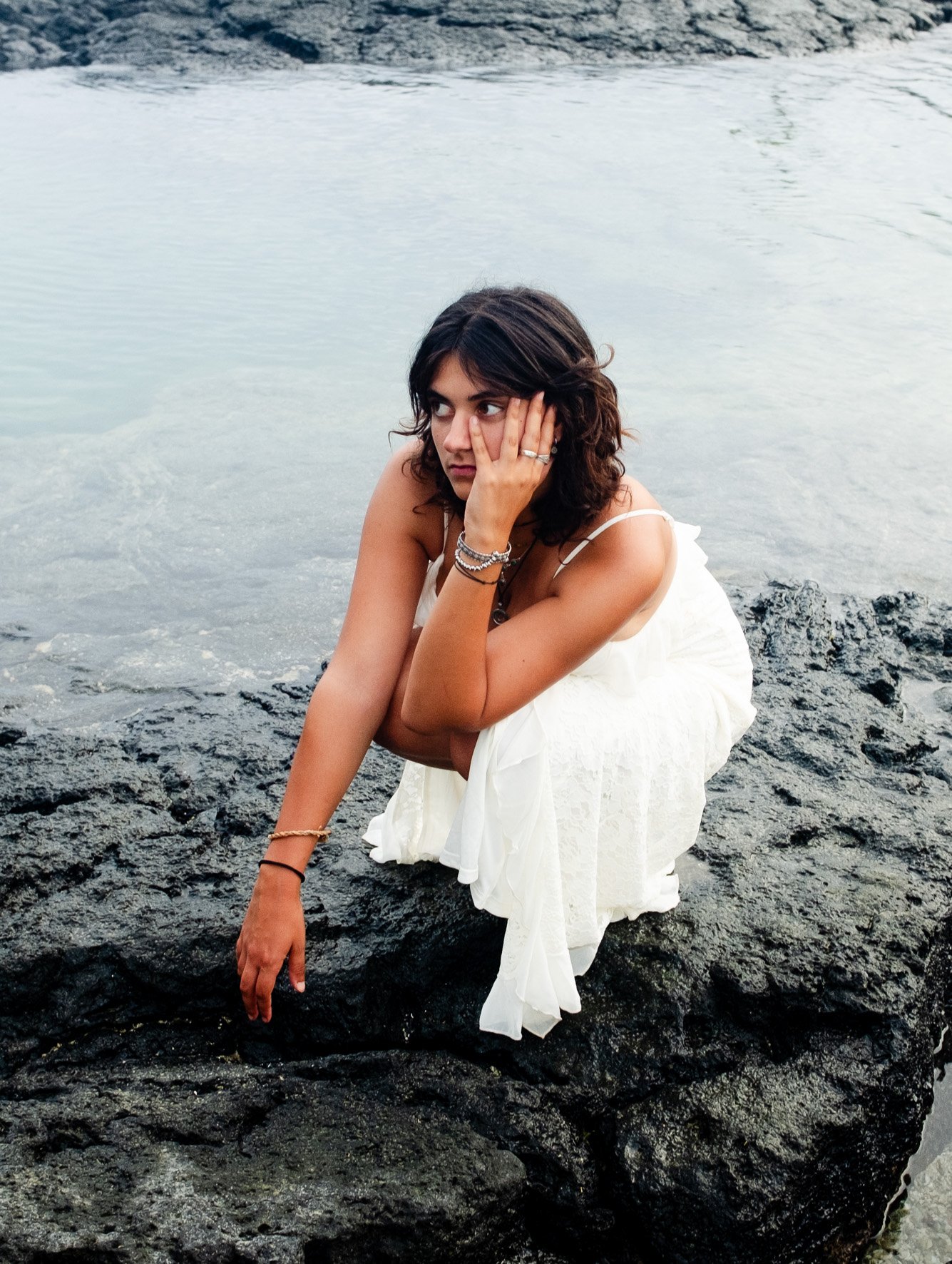 Jeune femme assise sur des rochers près de l'eau, portant une robe blanche, regardant pensivement avec sa main sur la joue.