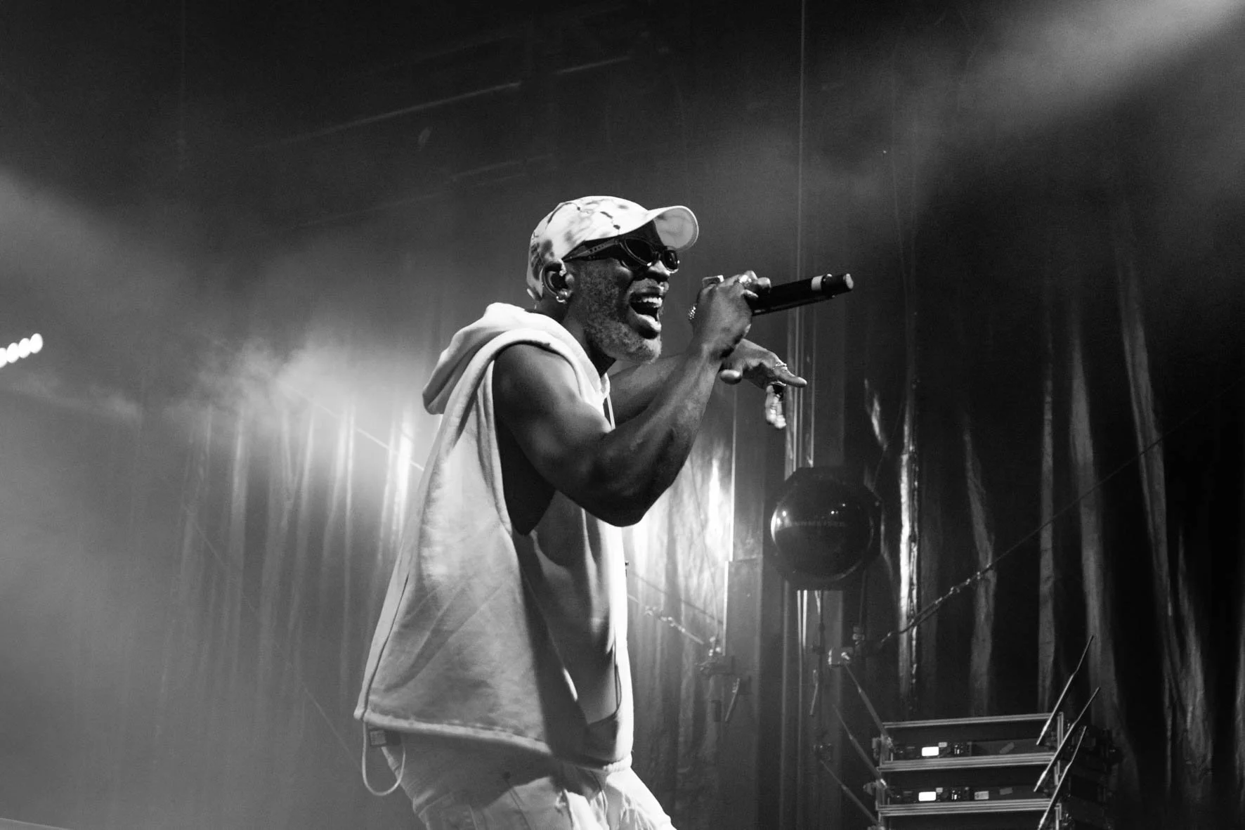 Rad cartier (rappeur) performant sur scène au week- end des curiosités. Il porte des lunettes de soleil, une casquette et un sweat à capuche. Photographie en noir et blanc.