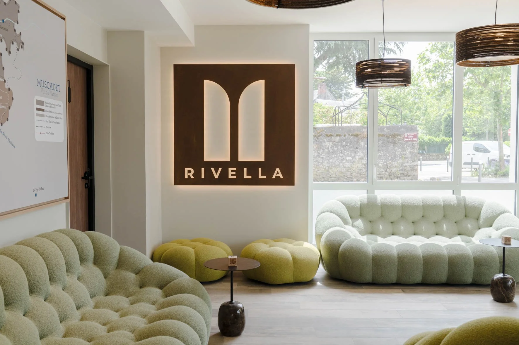 Salon d'accueil avec un grand canapé blanc en forme de boucle, un mur avec un logo lumineux 'Rivella', des suspensions noires rondes au plafond, et de grandes fenêtres laissant entrer la lumière naturelle.