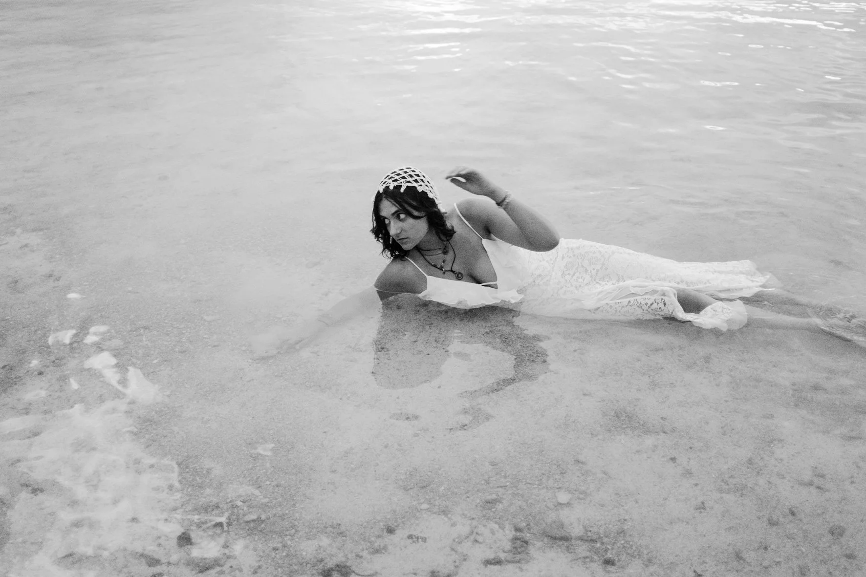 Une femme en robe blanche allongée dans l'eau de la mer, portant un chapeau en filet et des bijoux, avec un regard sérieux.