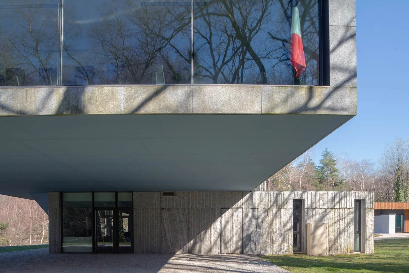 Vue d'une bâtiment moderne en béton avec un balcon en verre, un drapeau italien visible dans la fenêtre, et des arbres en arrière-plan.