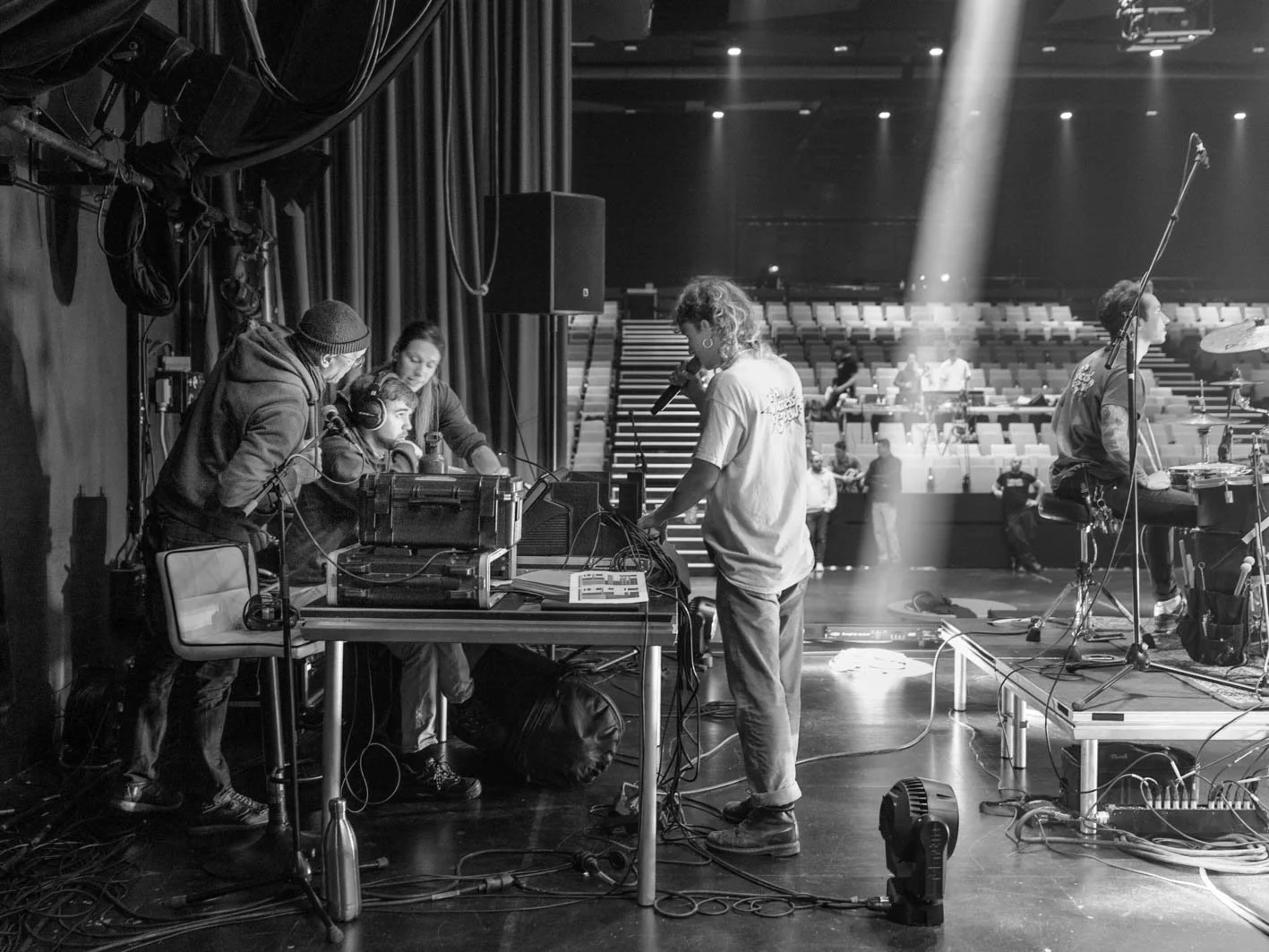 Une équipe technique en préparation sur scène avec un chanteur à microphone lors d'une répétition ou concert en salle de spectacle vide, en noir et blanc.