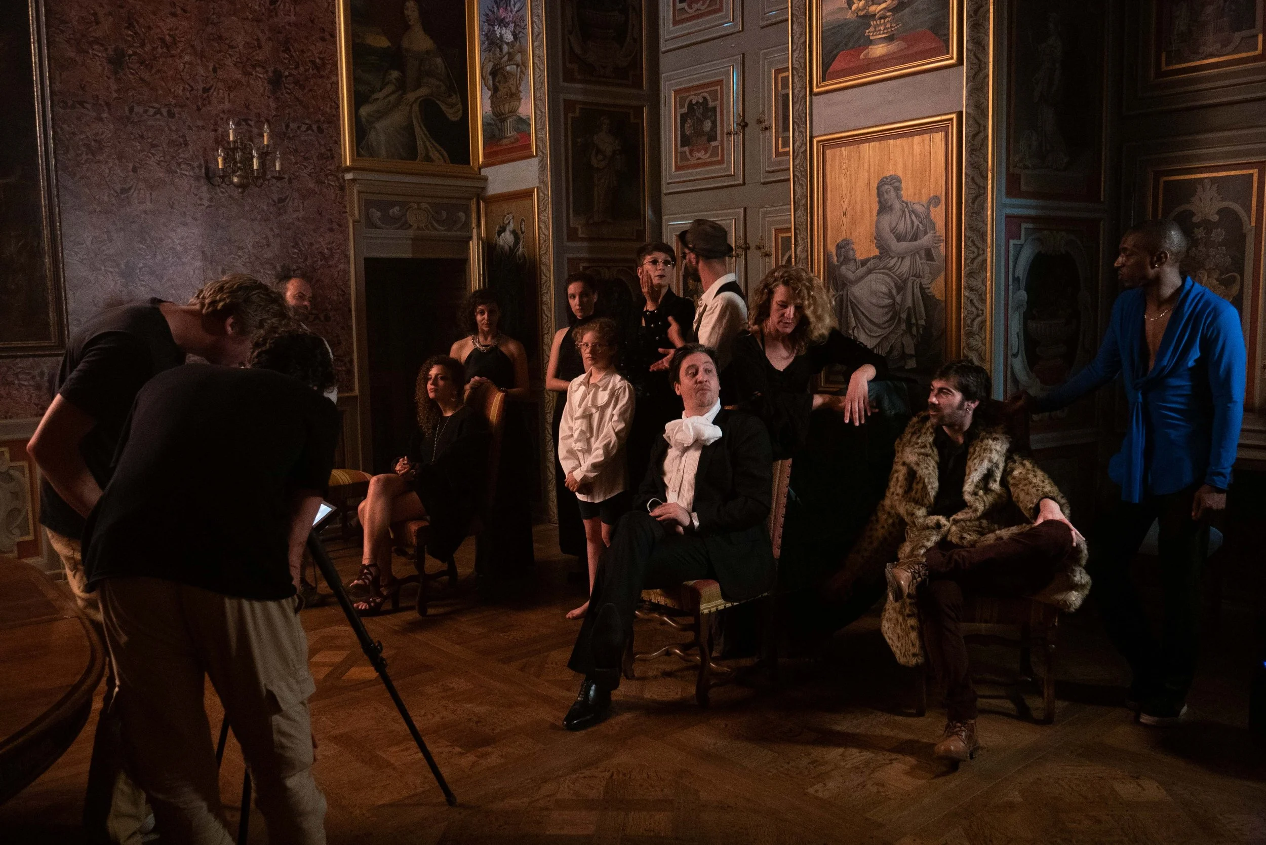 Groupe de personnes dans une salle ornée de tableaux et de moulures dorées, en train de discuter ou d'attendre.