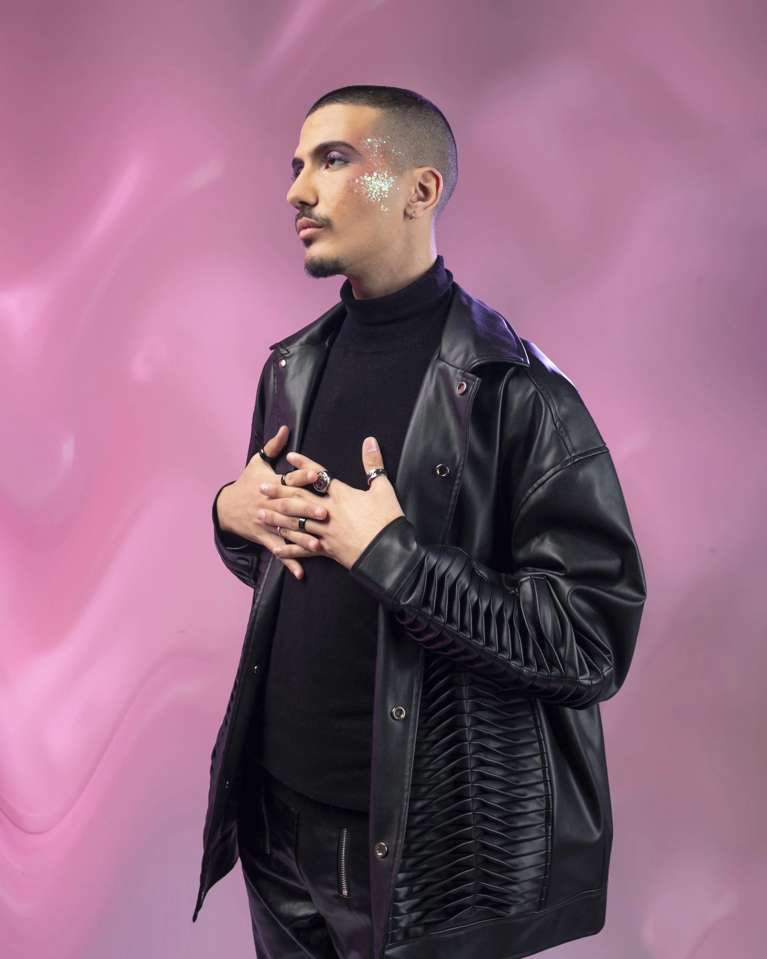 Portrait d'un homme avec maquillage violet, bijoux, portant une veste en cuir noir, posé devant un fond rose flou.