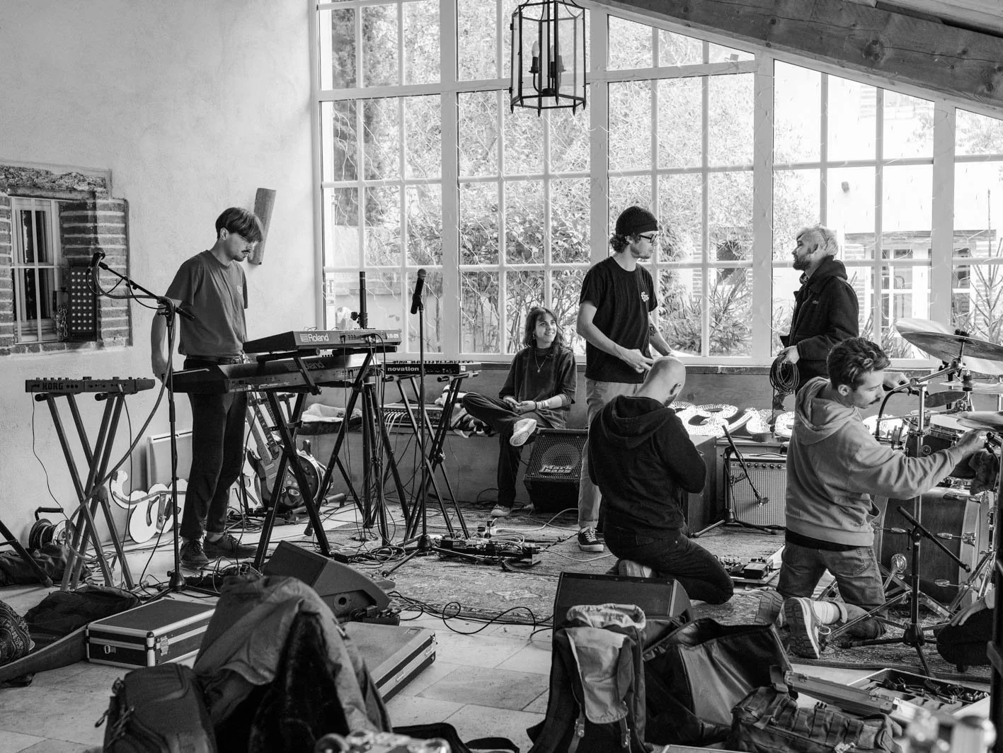 Un groupe de musiciens en répétition dans une salle lumineuse avec de grandes fenêtres. On voit plusieurs instruments électroniques, guitares, batteries, microphones et câbles partout. Le groupe semble discuter et préparer leur performance.