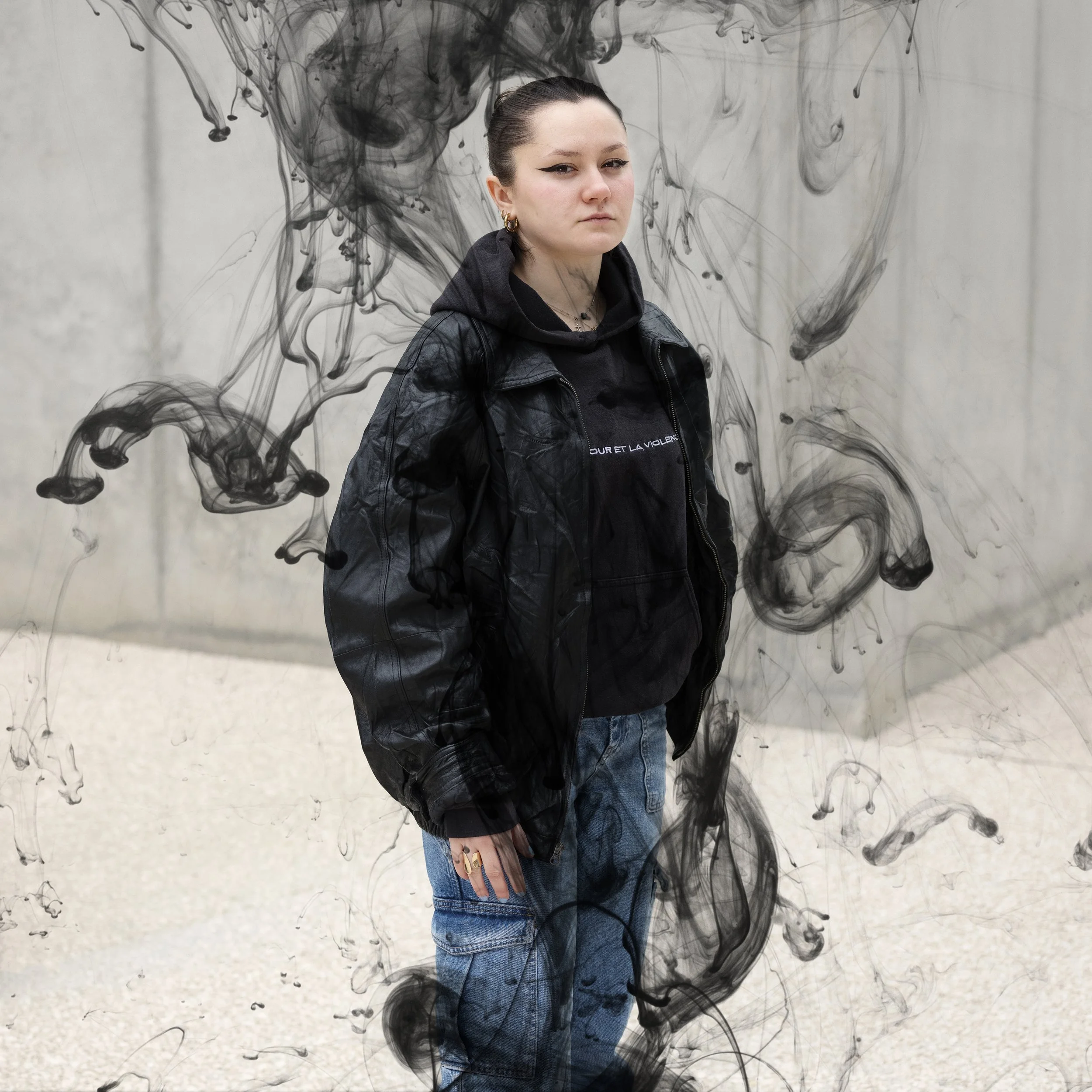 Jeune femme portant une veste en cuir, un sweat à capuche noire et un jean, debout devant une œuvre d'art en noir et blanc avec des motifs d'encre en mouvement.