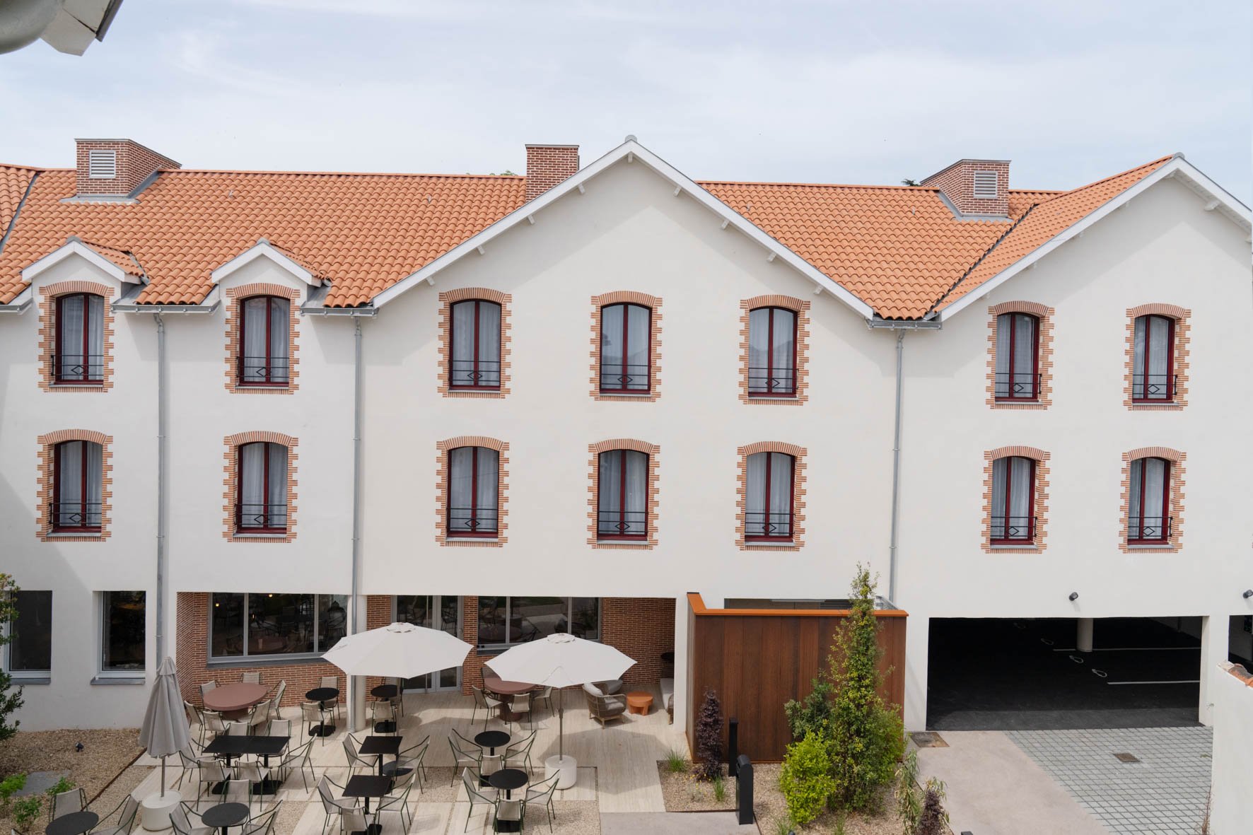 Un bâtiment résidentiel avec un toit en tuiles orange et des fenêtres à encadrements en briques rouges. Il y a une terrasse avec des tables et des parasols devant l'immeuble.