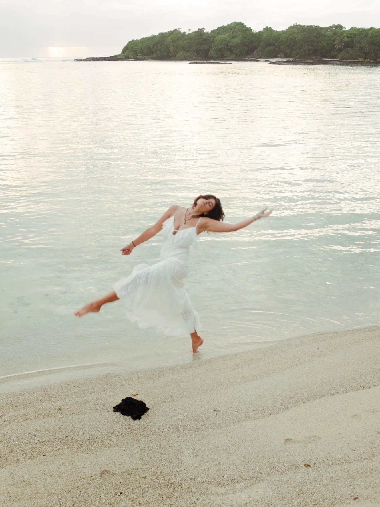 Une femme en robe blanche danse dans l'eau près de la plage, avec un paysage d'île verte en arrière-plan.