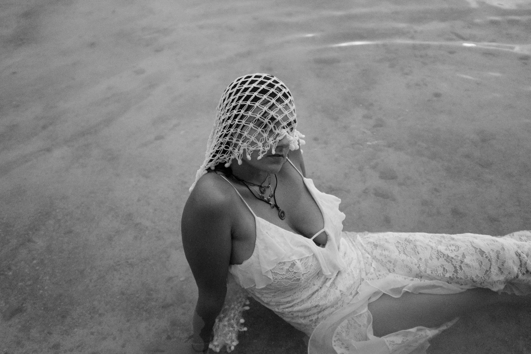 Une femme en robe blanche allongée sur la plage, portant un filet de pêche sur la tête, en noir et blanc.