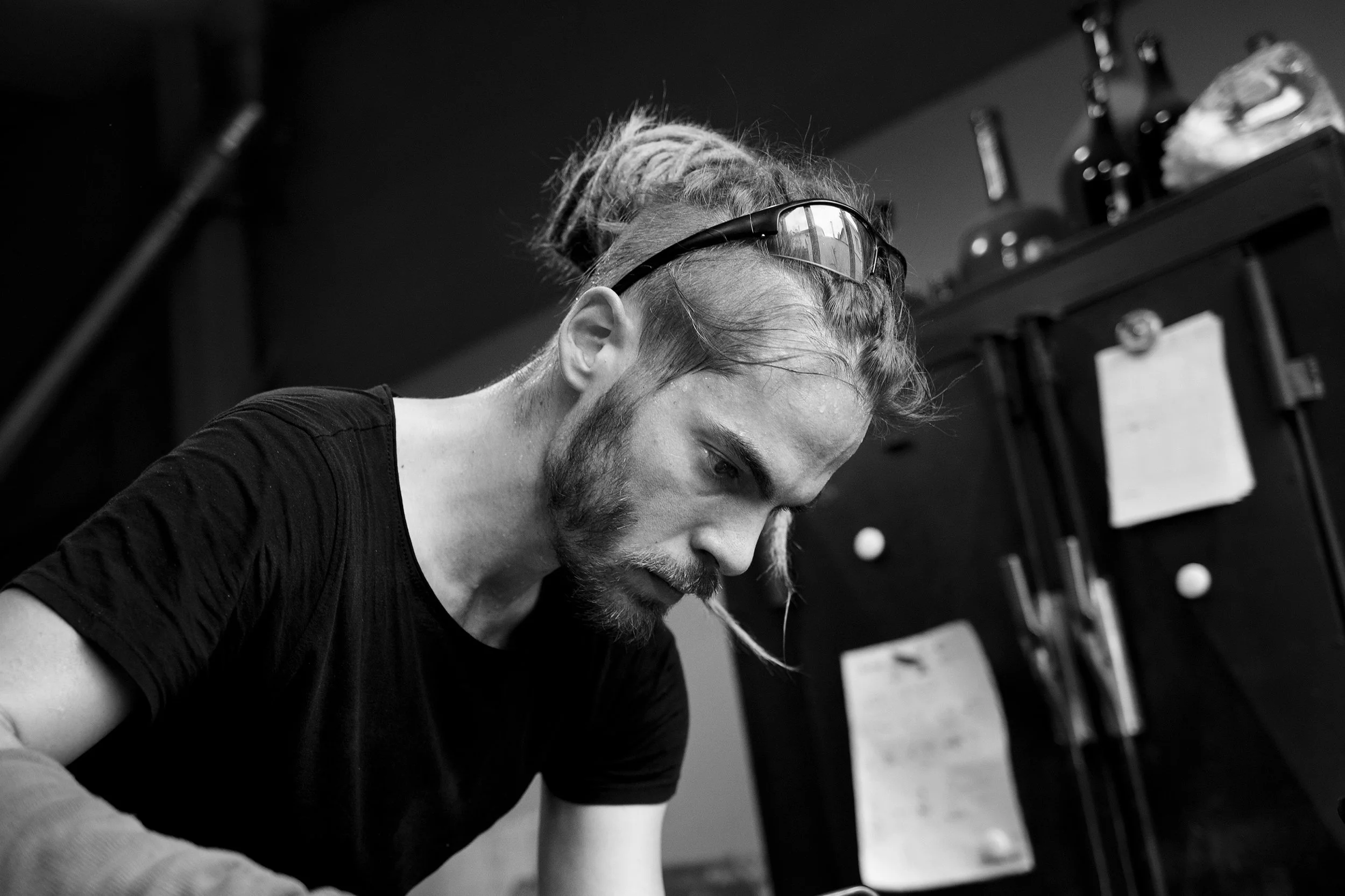 Un homme avec des dreads, portant des lunettes de soleil sur sa tête, regarde vers le bas dans un environnement intérieur, probablement un studio ou un espace de travail.