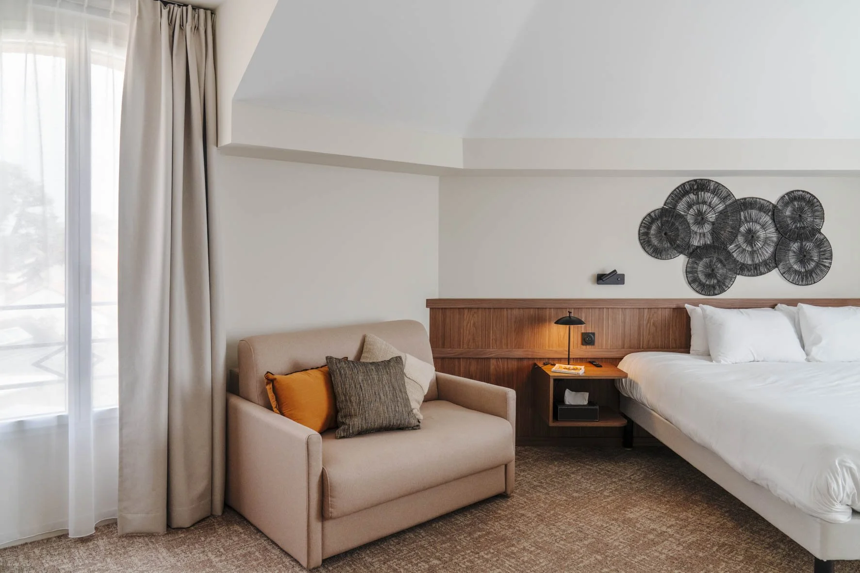 Chambre d'hôtel avec un lit blanc, un panneau en bois au mur, une décoration murale en forme de cercles noirs, une lampe de chevet et un petit canapé beige avec des coussins