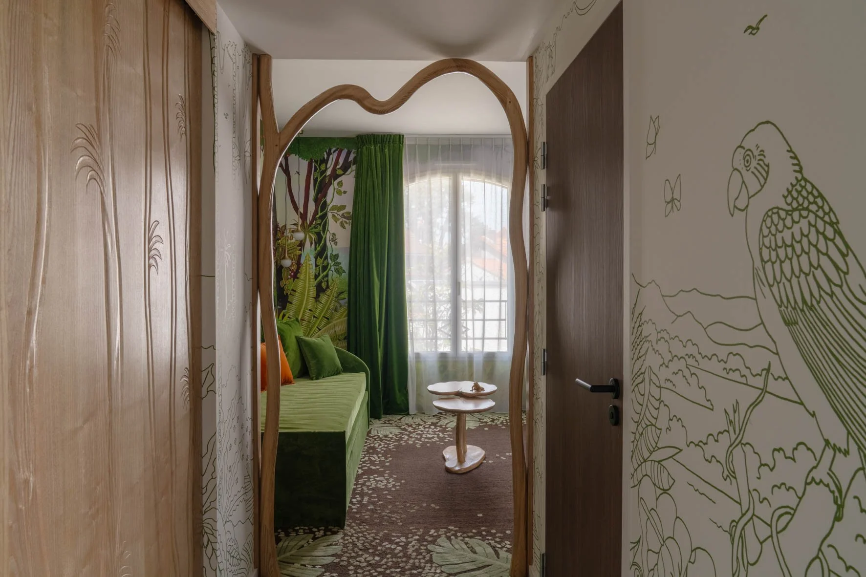 Une chambre avec un décor de jungle, comprenant un canapé vert, des voilages transparents, un mur avec une fresque de perroquet et de nature, et un miroir en bois avec une forme organique.