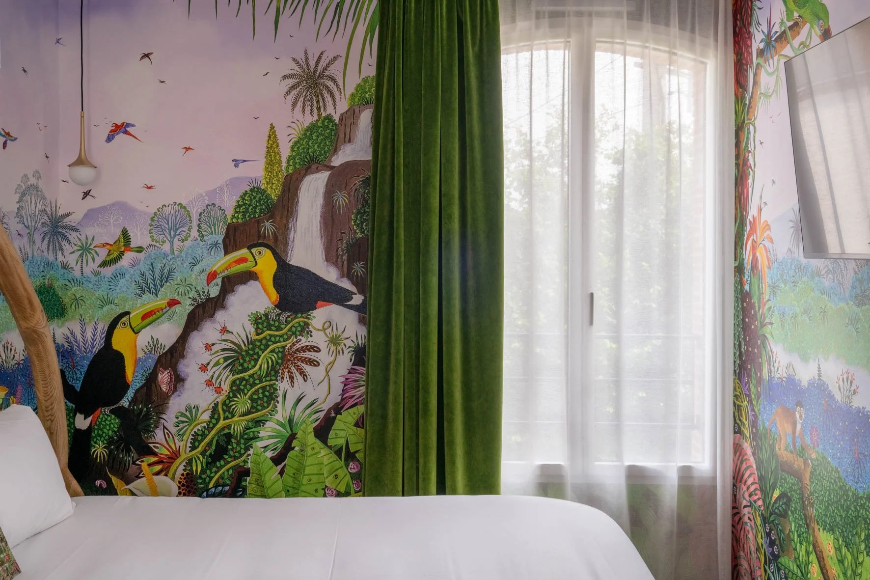 Mur peinte avec un paysage tropical, y compris des toucans, des cascades, des palmiers et des oiseaux, derrière des rideaux verts et une fenêtre avec volets.
