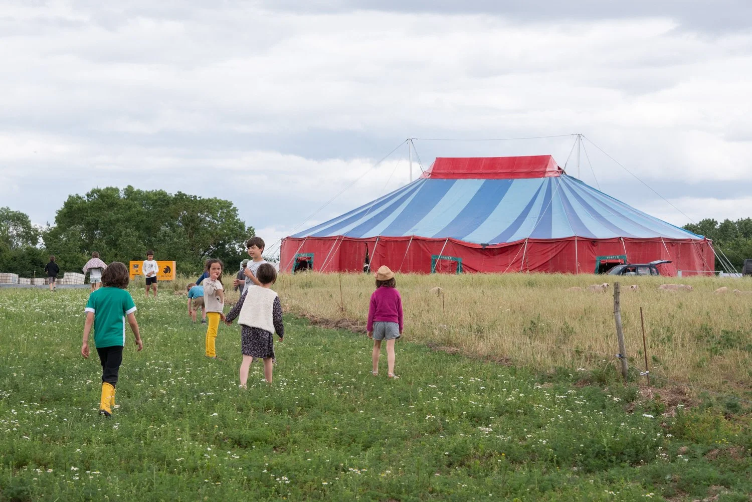 Un groupe d'enfants jouent dans un champ vert près d'un grand chapiteau de cirque rouge et bleu, avec des arbres en arrière-plan et un ciel nuageux.