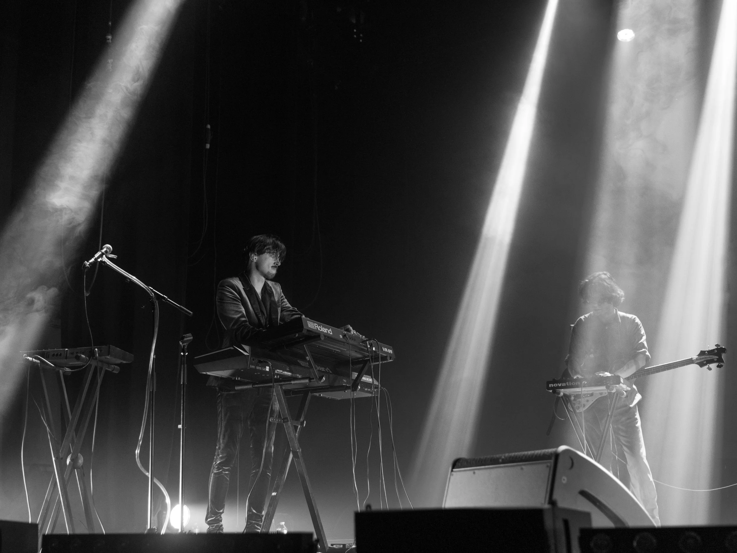 Deux musiciens jouent sur scène, éclairés par des faisceaux de lumière. Le premier à gauche, avec une guitare, et le second au centre utilisant un clavier. Scène sombre avec un micro visible à côté du guitariste.
