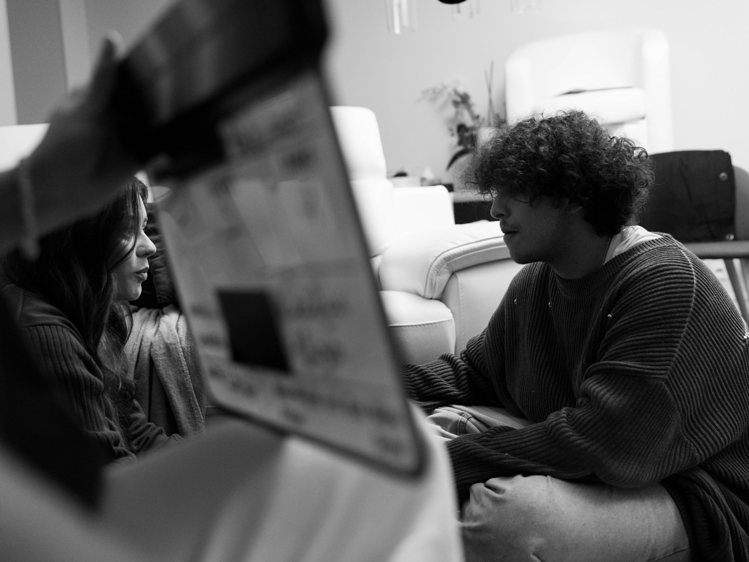 Deux personnes assises au sol séparé par un clap de tournage au premier plan. Une femme avec des cheveux longs et une autre personne avec des cheveux bouclés, échanges dans un environnement intérieur. La photo est en noir et blanc.