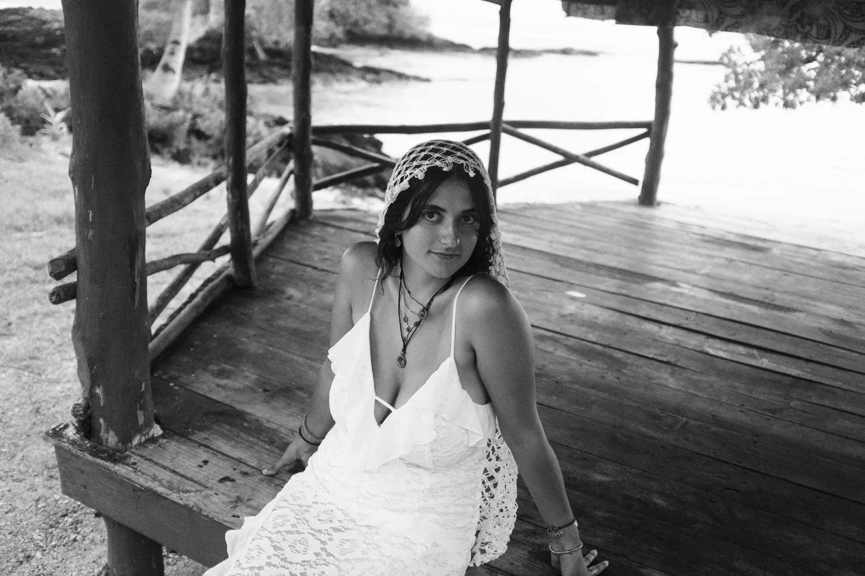 Une jeune femme en robe blanche et un chapeau en crochet posé sur une structure en bois, près d'un plan d'eau, prise en noir et blanc.